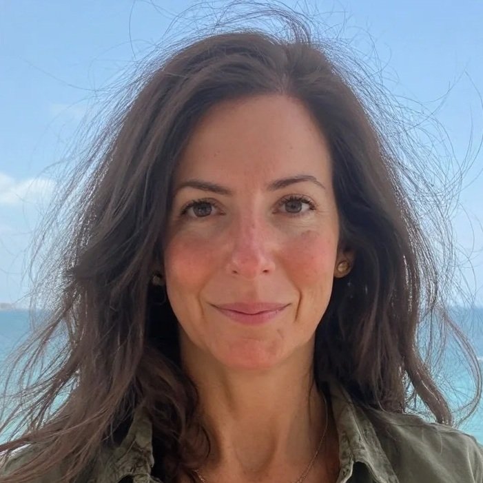 一位长发乌黑的女子在白天的户外微笑，背景是蔚蓝的天空与海洋。