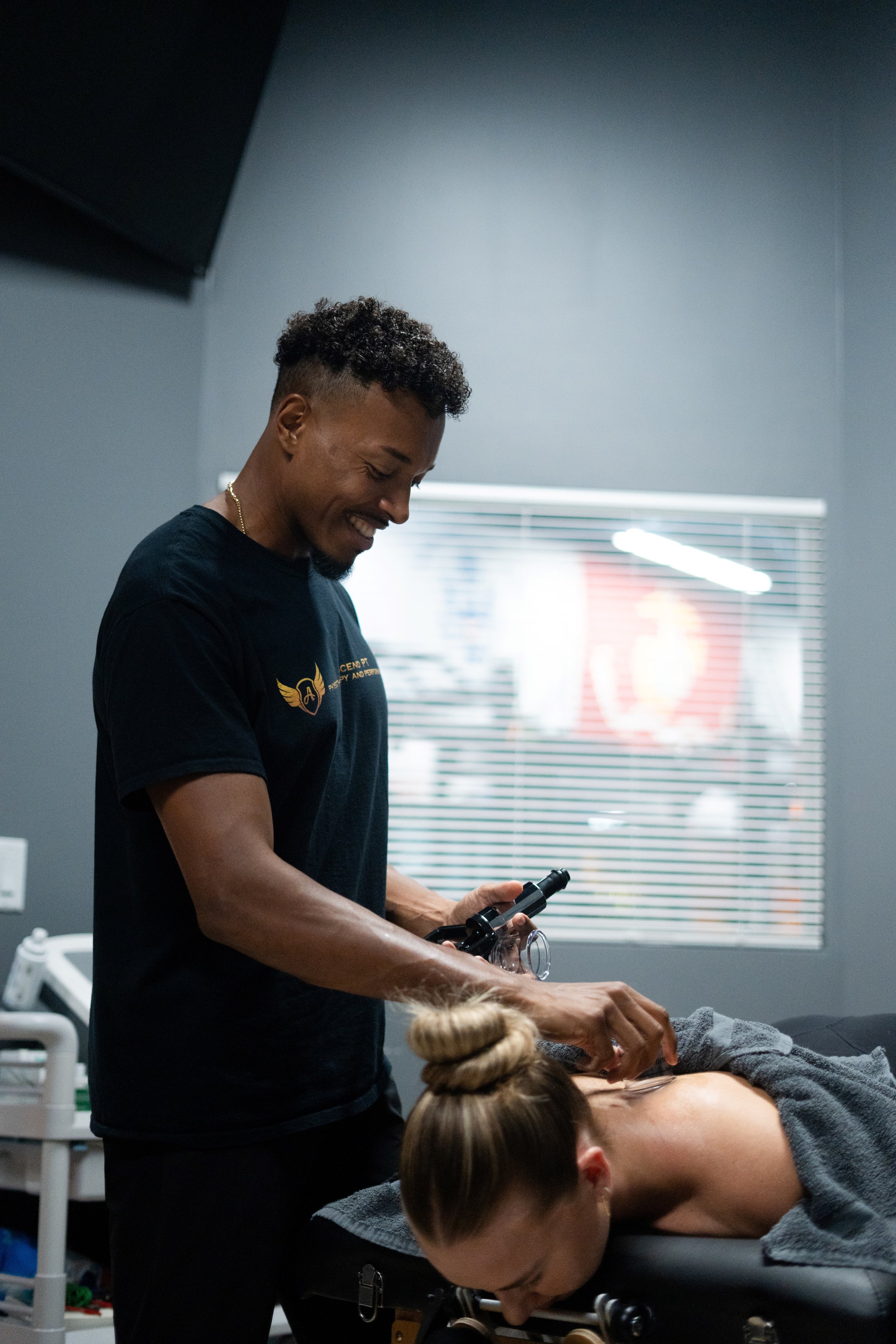A man with curly hair and a black shirt is giving a massage or therapy to a woman lying face down on a treatment table. The setting appears to be a wellness or massage clinic, with a window with blinds and various medical or massage equipment in the background.