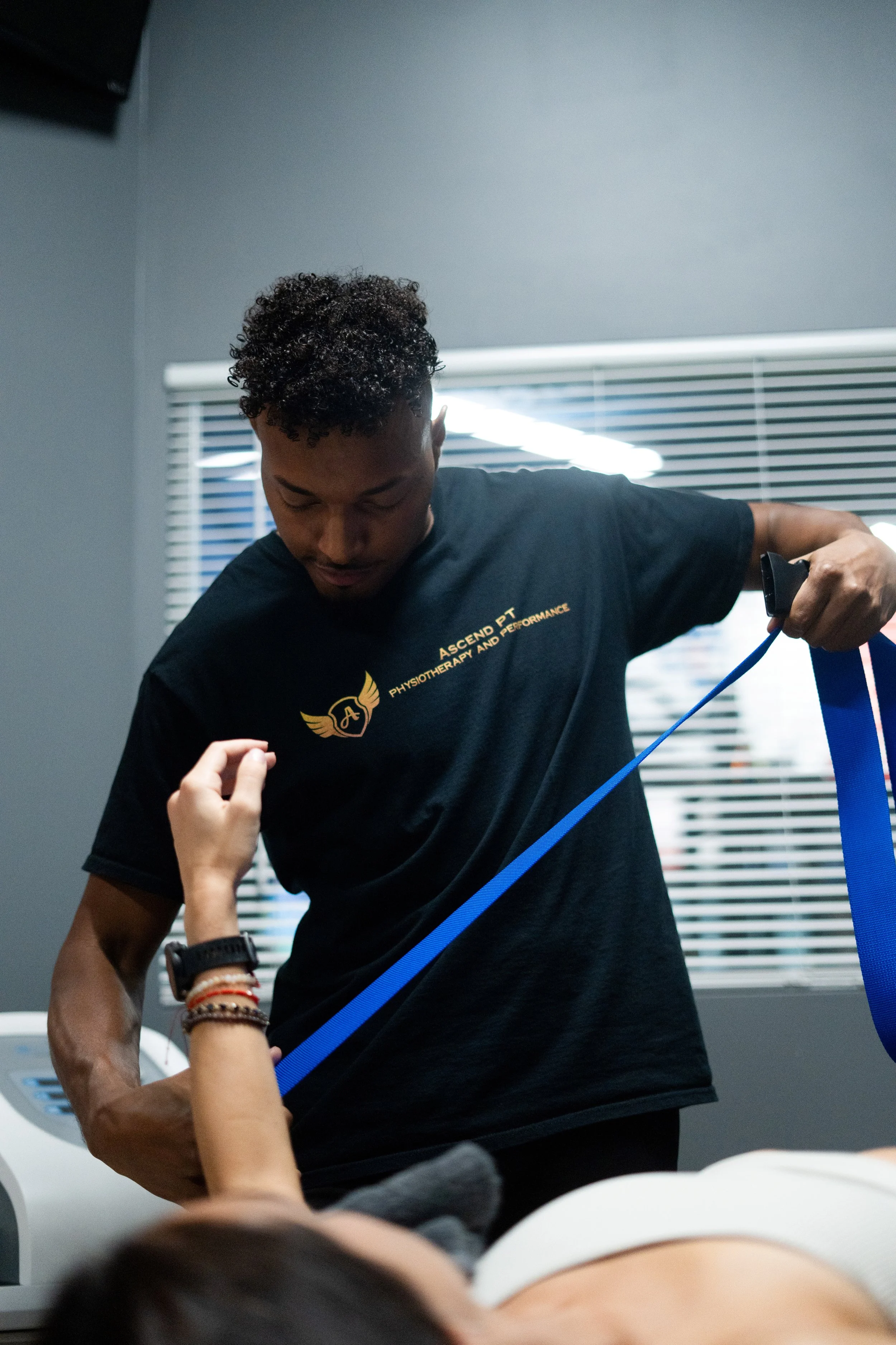 A healthcare professional or physical therapist helps a patient with resistance band exercises in a clinical setting, with window blinds in the background.