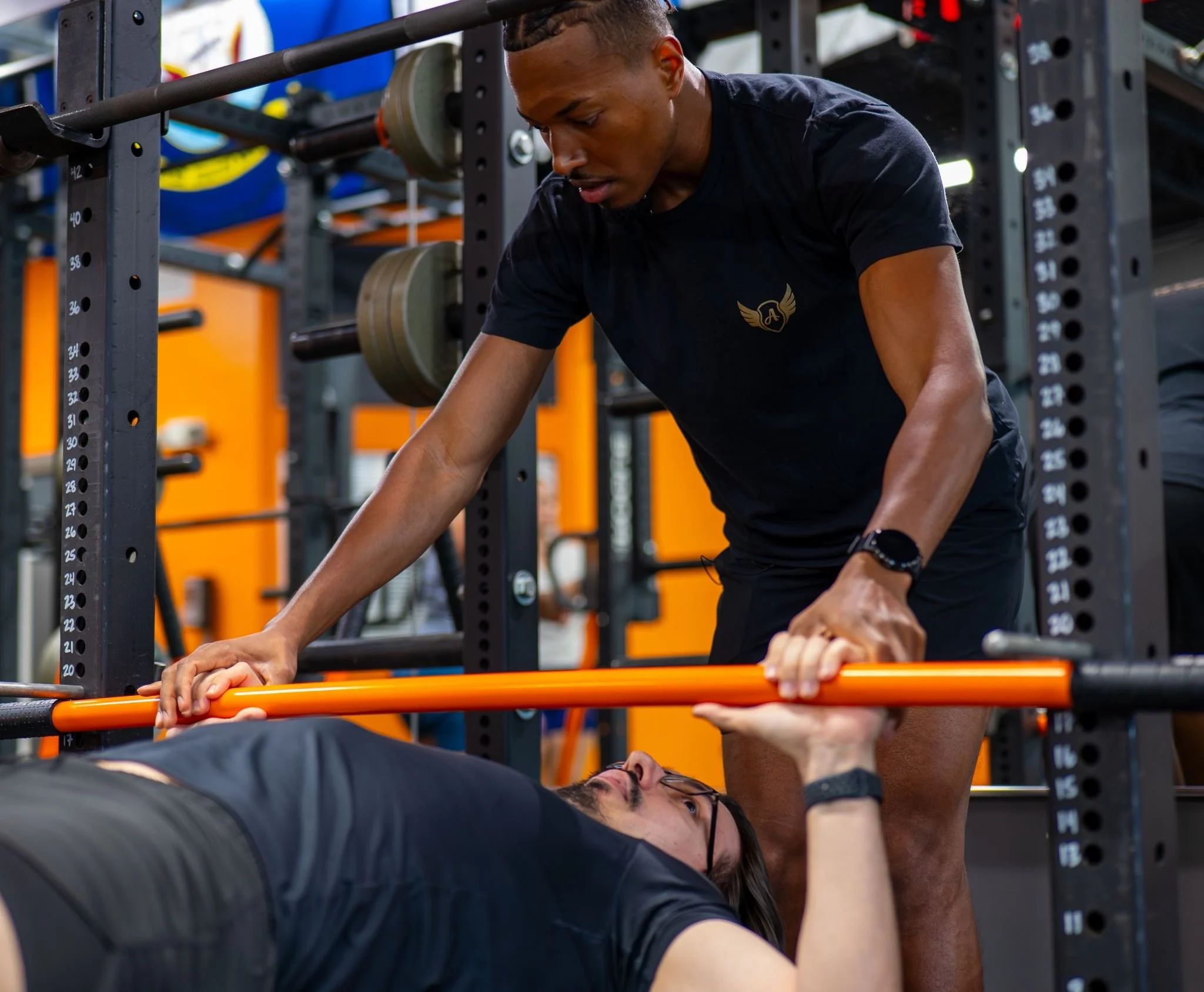 A man in a black T-shirt and glasses lying on a weight bench and lifting weights with a woman in a black shirt assisting him at a gym with exercise equipment in the background.