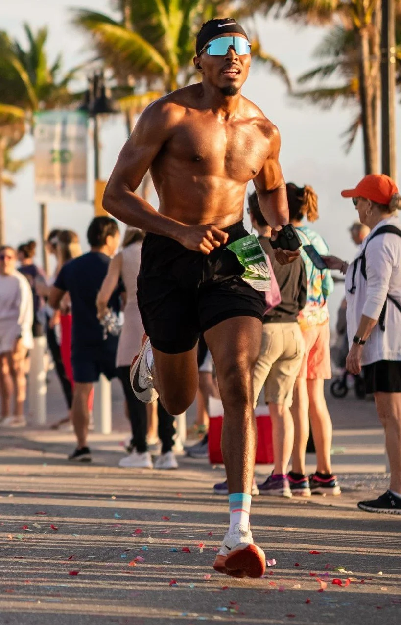A shirtless male runner with sunglasses and a headband participating in a race near palm trees, with a crowd of spectators and other runners in the background.