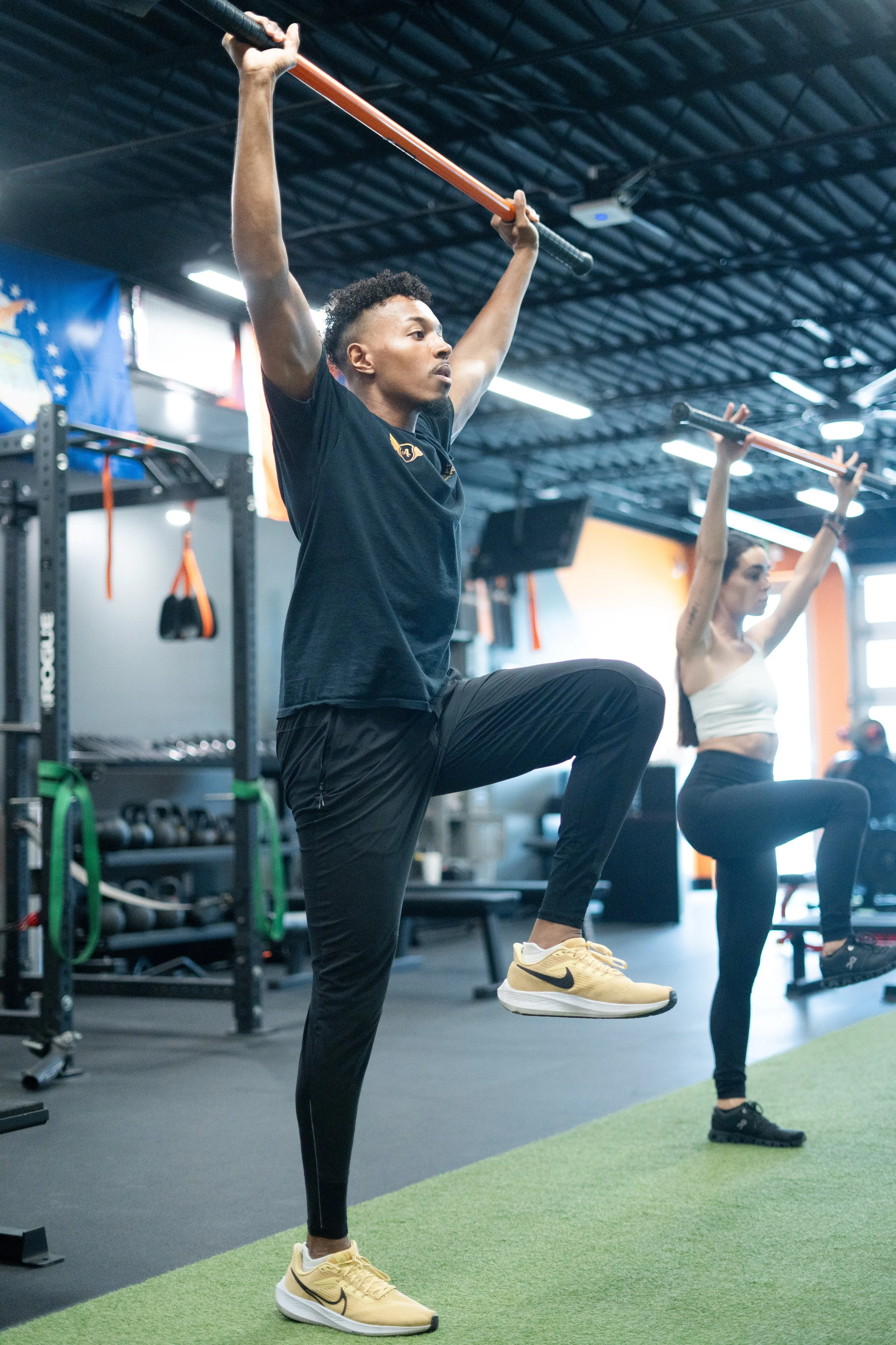 A man and a woman exercising in a gym, lifting weights overhead, standing on one leg with one knee bent, wearing athletic clothing.