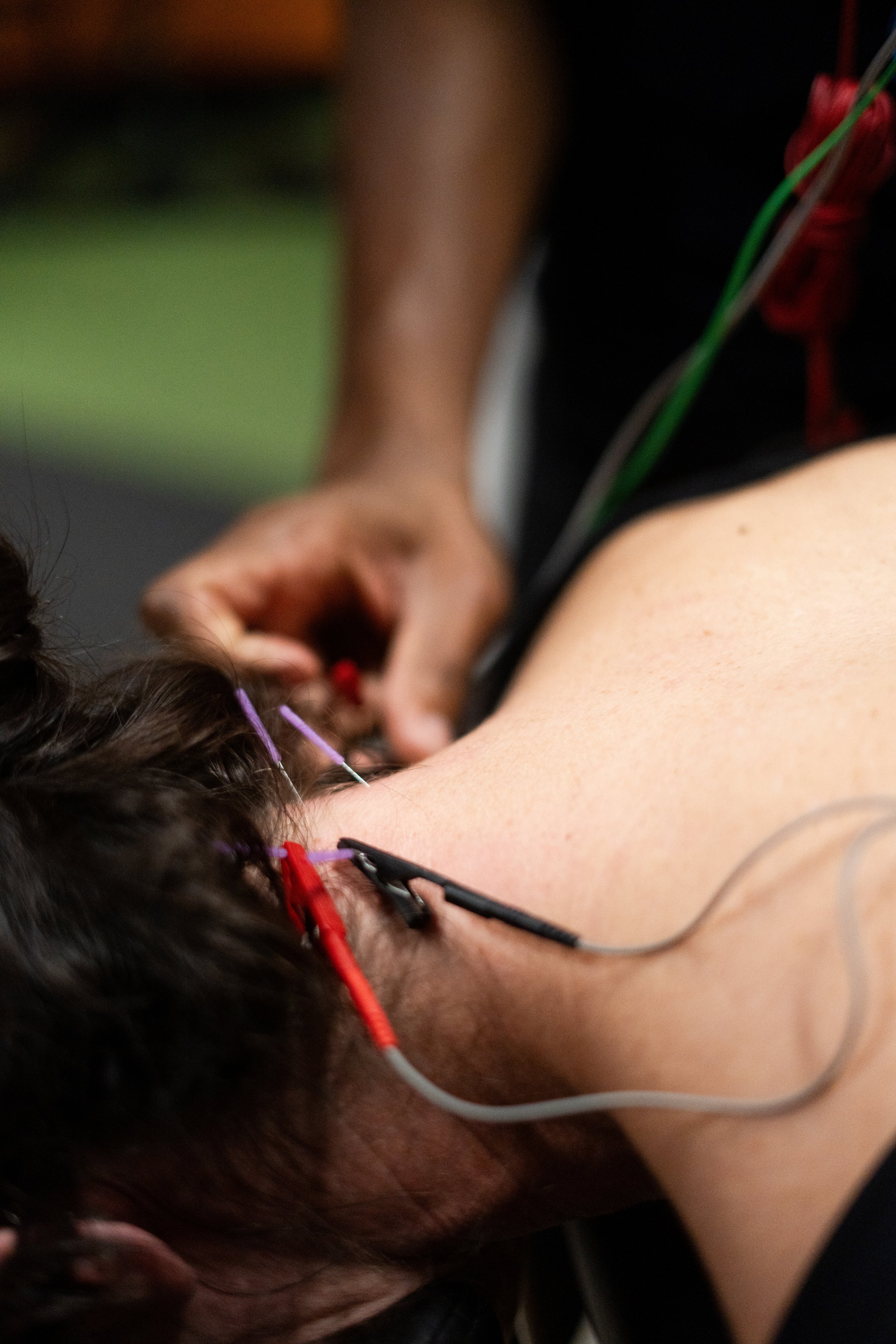 Close-up photo of a person lying down with their head turned to the side, showing a portion of their neck and ear. The person has dark hair, and there are colorful earbuds in their ear. The focus is on the person's neck and head, with the background blurred.