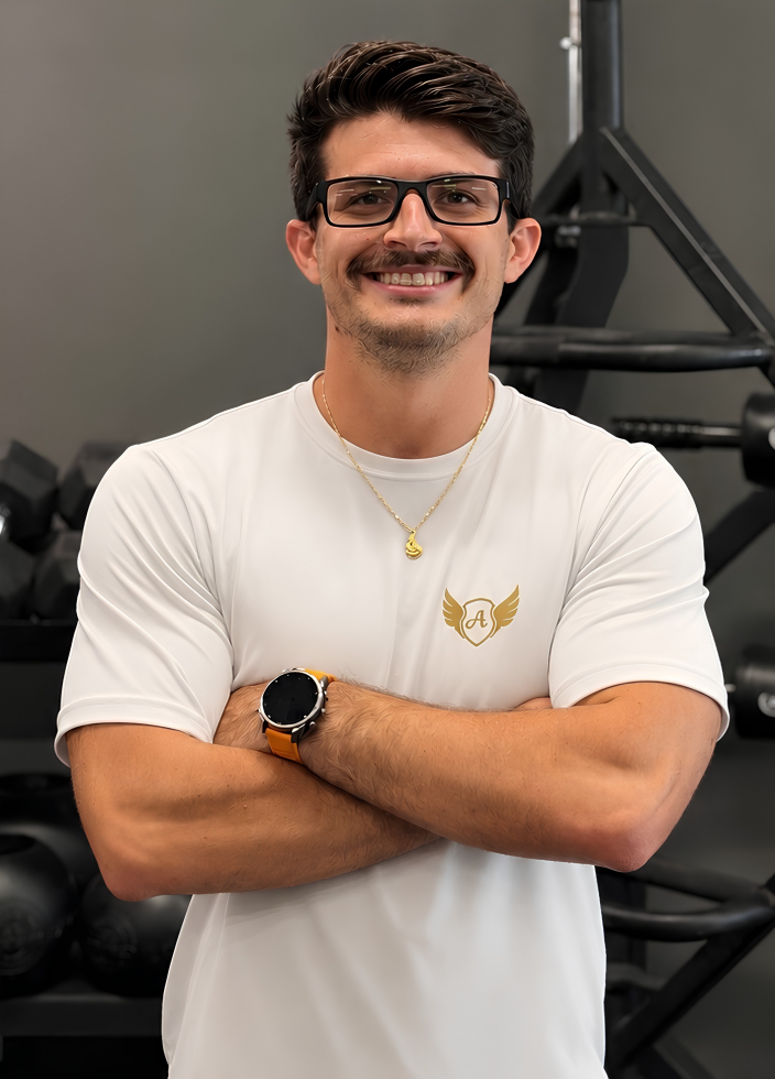 A young man with dark hair, beard, and glasses, smiling with arms crossed in a gym, wearing a white t-shirt with a gold emblem, a gold necklace, and a smartwatch, with gym equipment in the background.