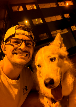 A smiling man wearing glasses and a baseball cap taking a selfie with a large, happy, fluffy dog at night outside a building with lit windows.