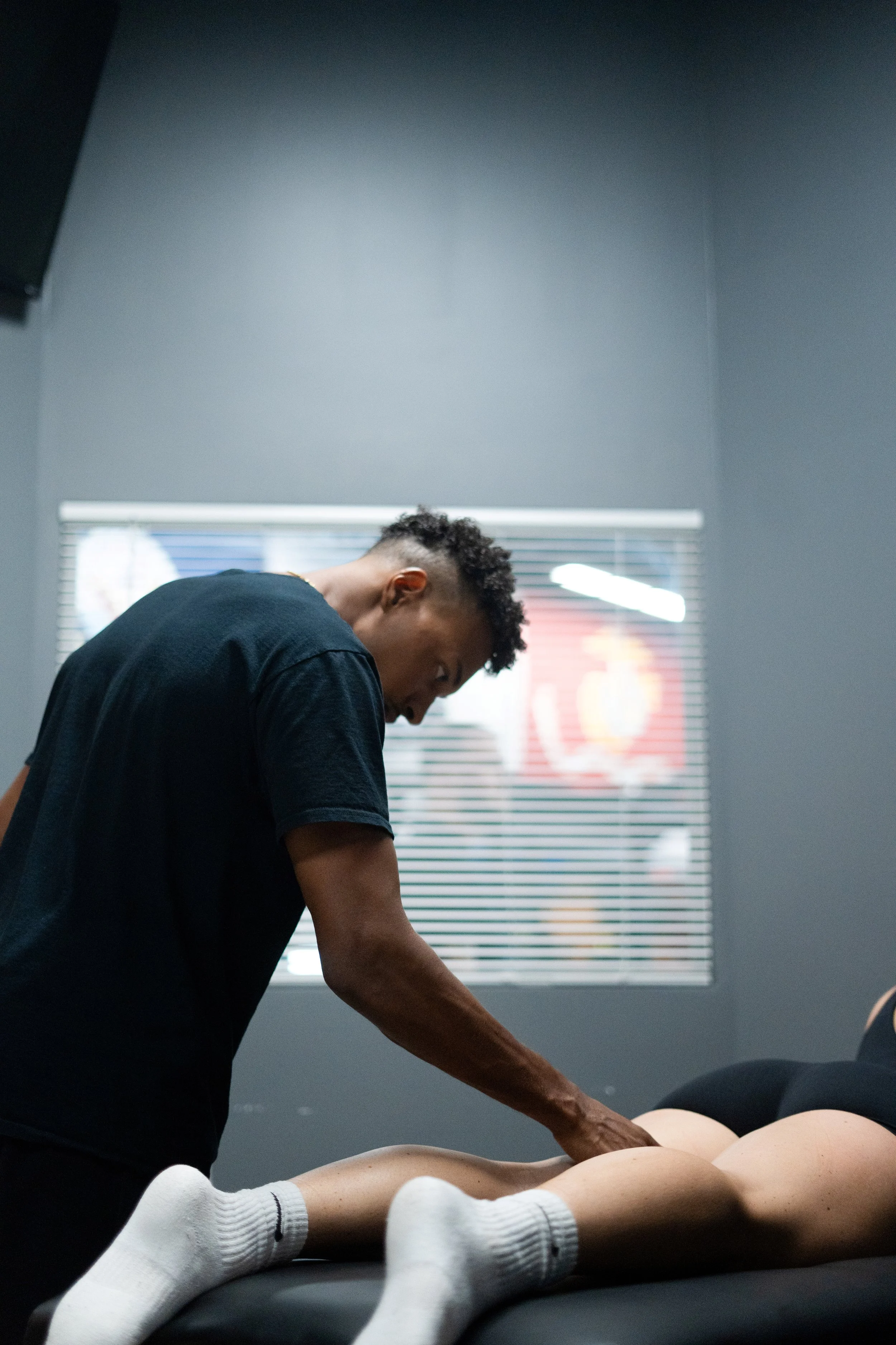 A person with short curly hair performing a massage or physical therapy on a mannequin or patient lying face down on a massage table, with room lighting coming through a window with blinds.