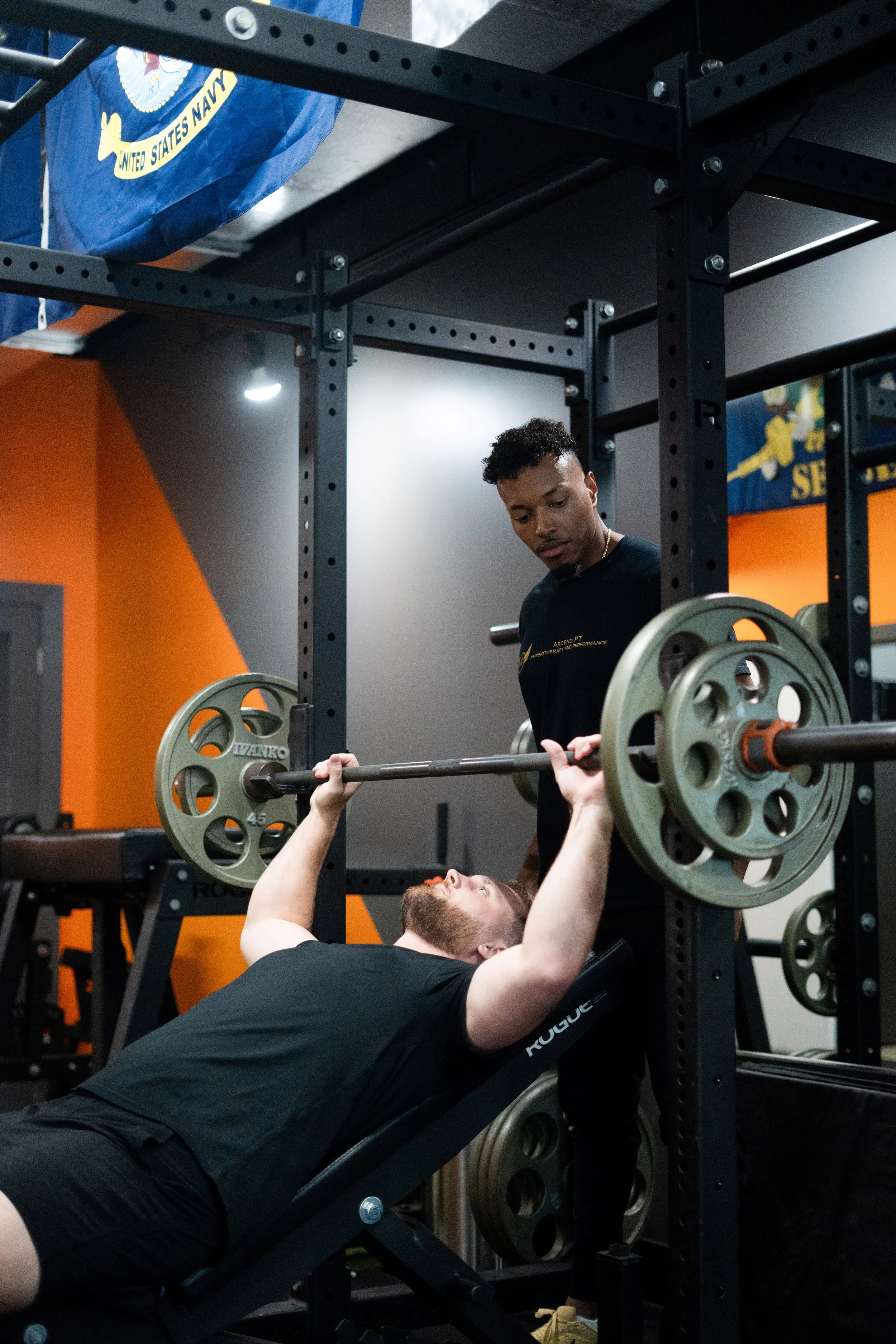 Person lying on a bench press during a workout while another person spots them. They are in a gym with orange and gray walls, and a US Navy flag is visible hanging from the ceiling.
