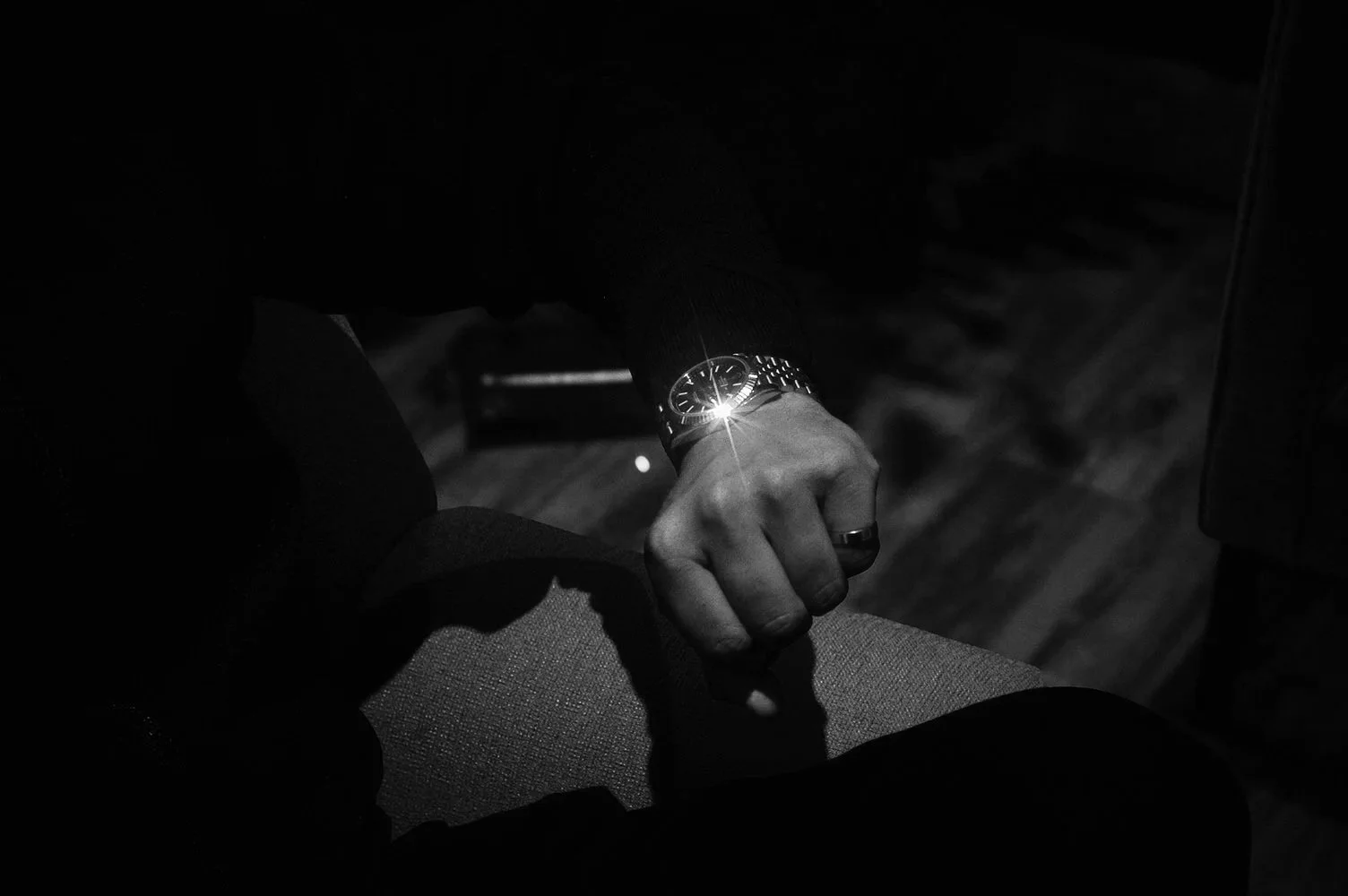 A person sitting with their hand resting on their knee, wearing a watch that reflects light, in a dark setting.