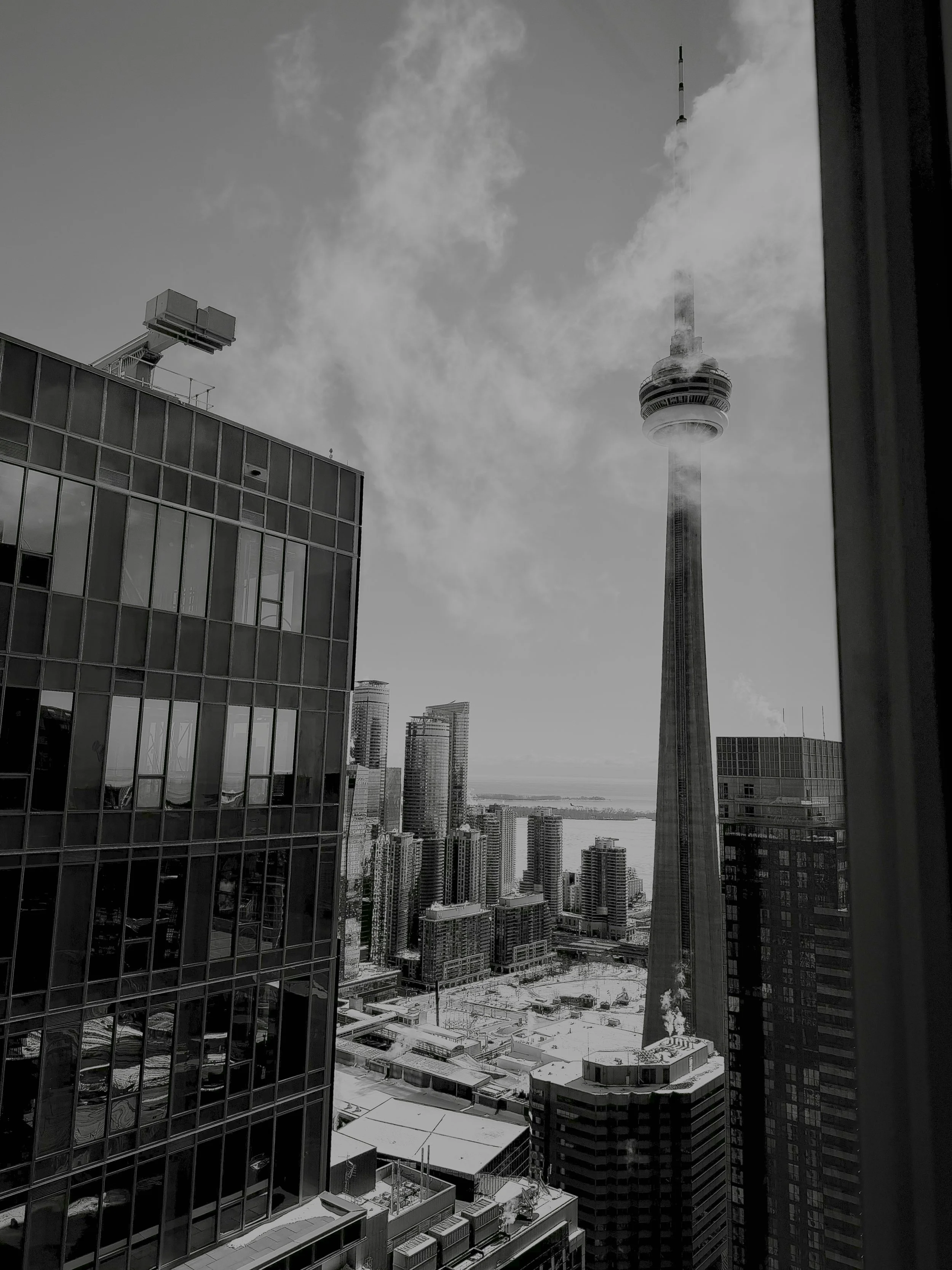A black and white photo of a city skyline showing tall buildings and CN Tower in Toronto, with clouds and smoke; viewed from a window.
