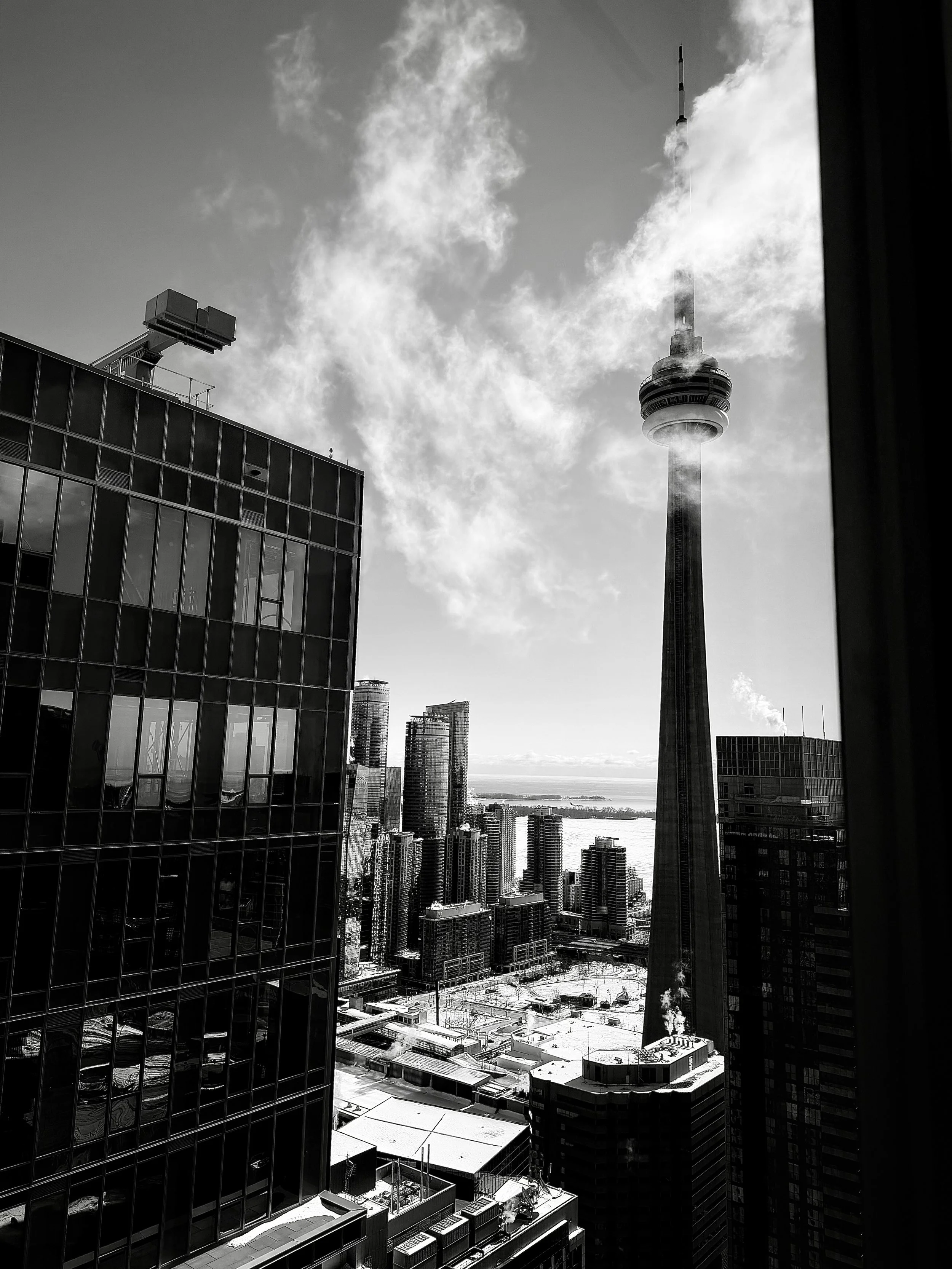 Black and white photo of a cityscape featuring the CN Tower in Toronto, with tall skyscrapers, some smoke or steam rising from a building, and a partly cloudy sky in the background.