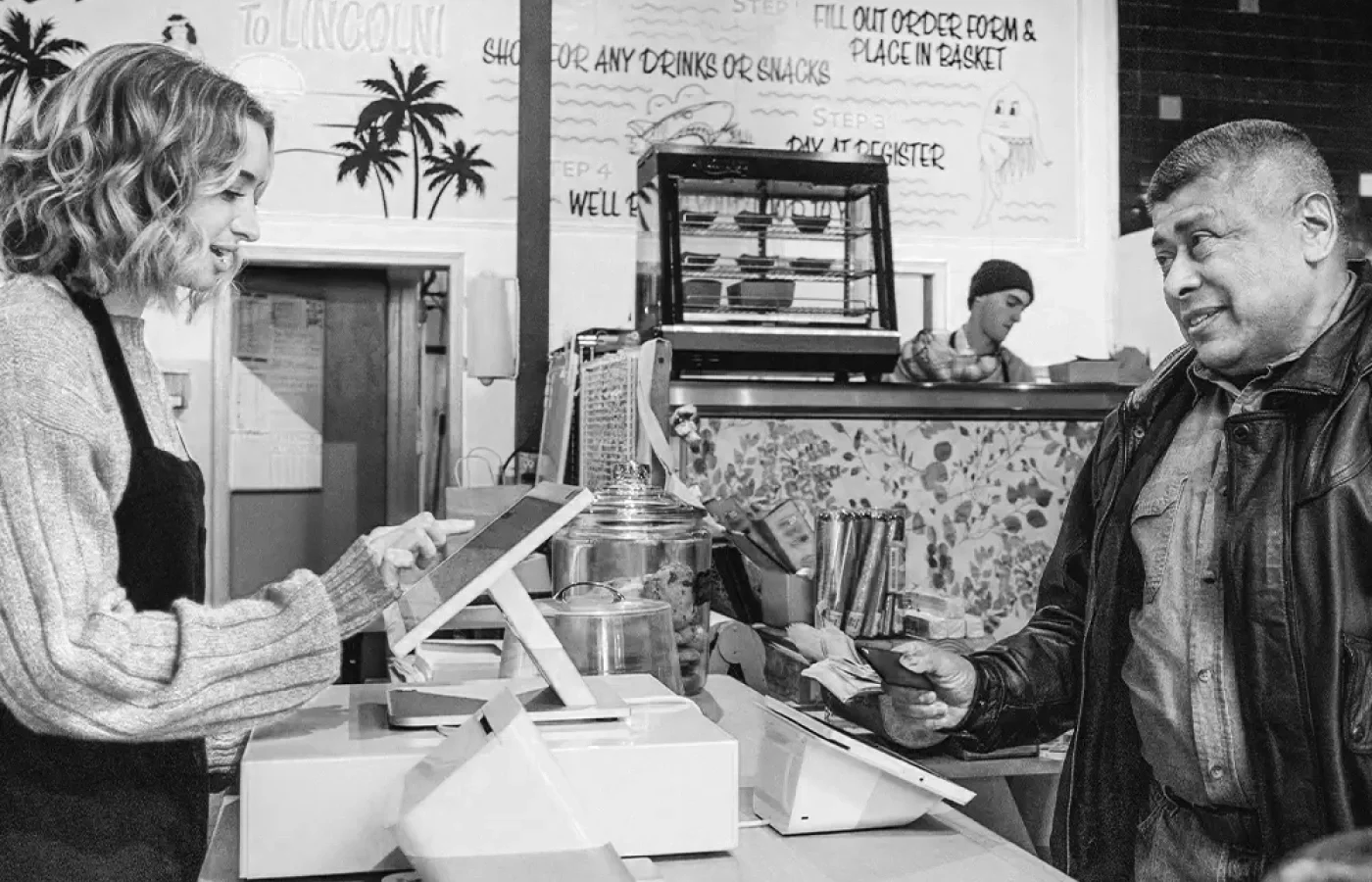 A woman and a man are standing at a counter in a cafe or restaurant, both looking at a digital device. The woman is on the left, with curly hair, wearing a sweater, and the man on the right, with short hair, wearing a leather jacket. There are jars and a cash register on the counter, with a striped tablecloth and a person in a beanie in the background.