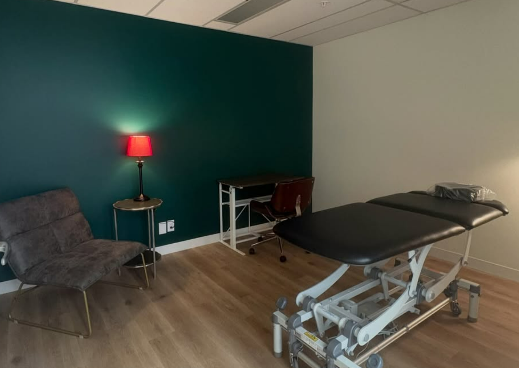 Medical examination room with examination table, a brown chair, a dark green wall, a red lamp on a side table, and wooden flooring.