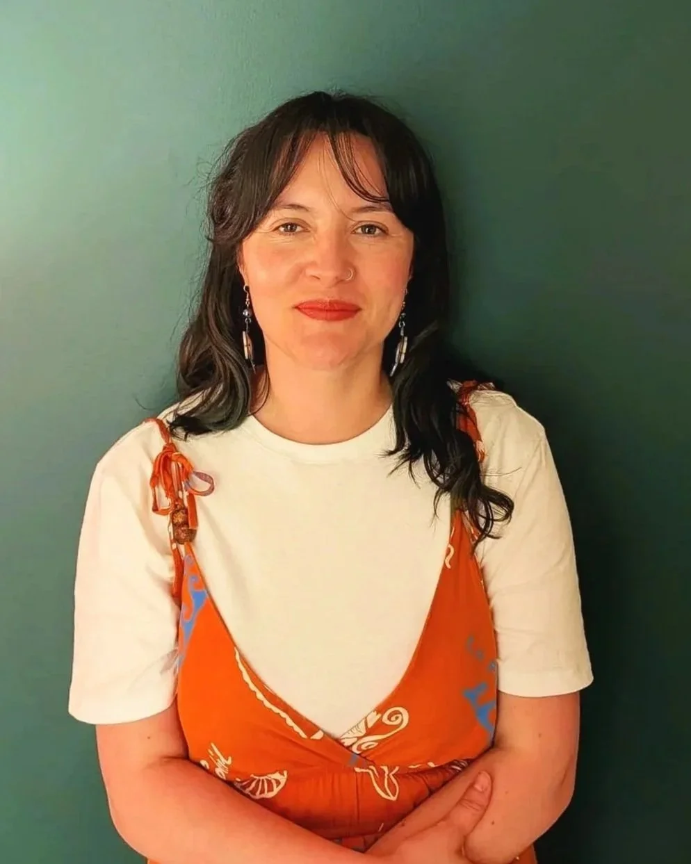 A woman with dark hair and earrings, standing against a green background, wearing a white t-shirt under an orange dress with swirl patterns.
