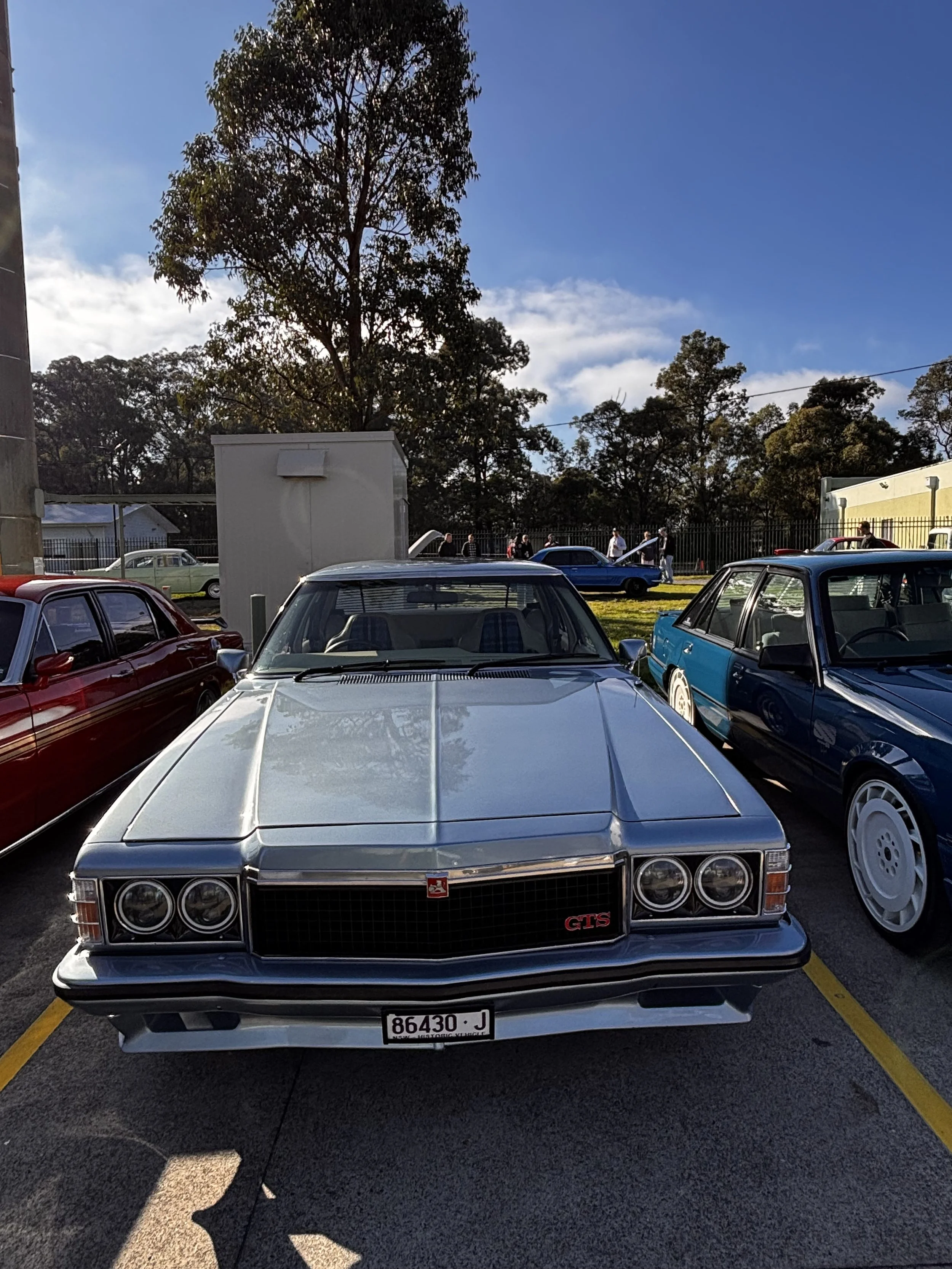 A classic silver Cadillac GTS parked in a parking lot with other vintage cars, trees, and a blue sky in the background.