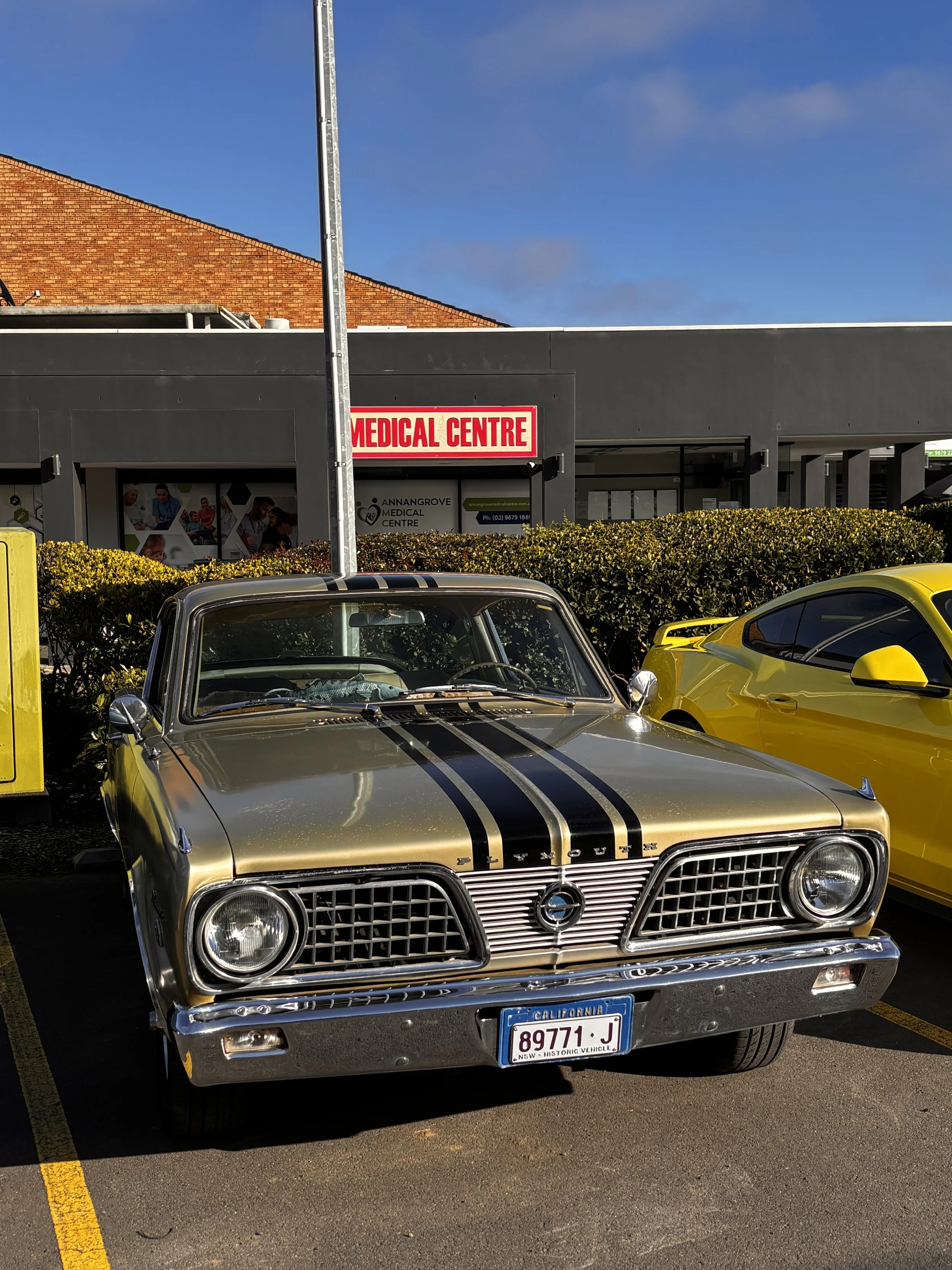 A vintage gold car with black racing stripes parked in a parking lot in front of a medical center. A yellow sports car is partially visible at the right edge of the image. The background includes a black building with a red and white sign that reads 