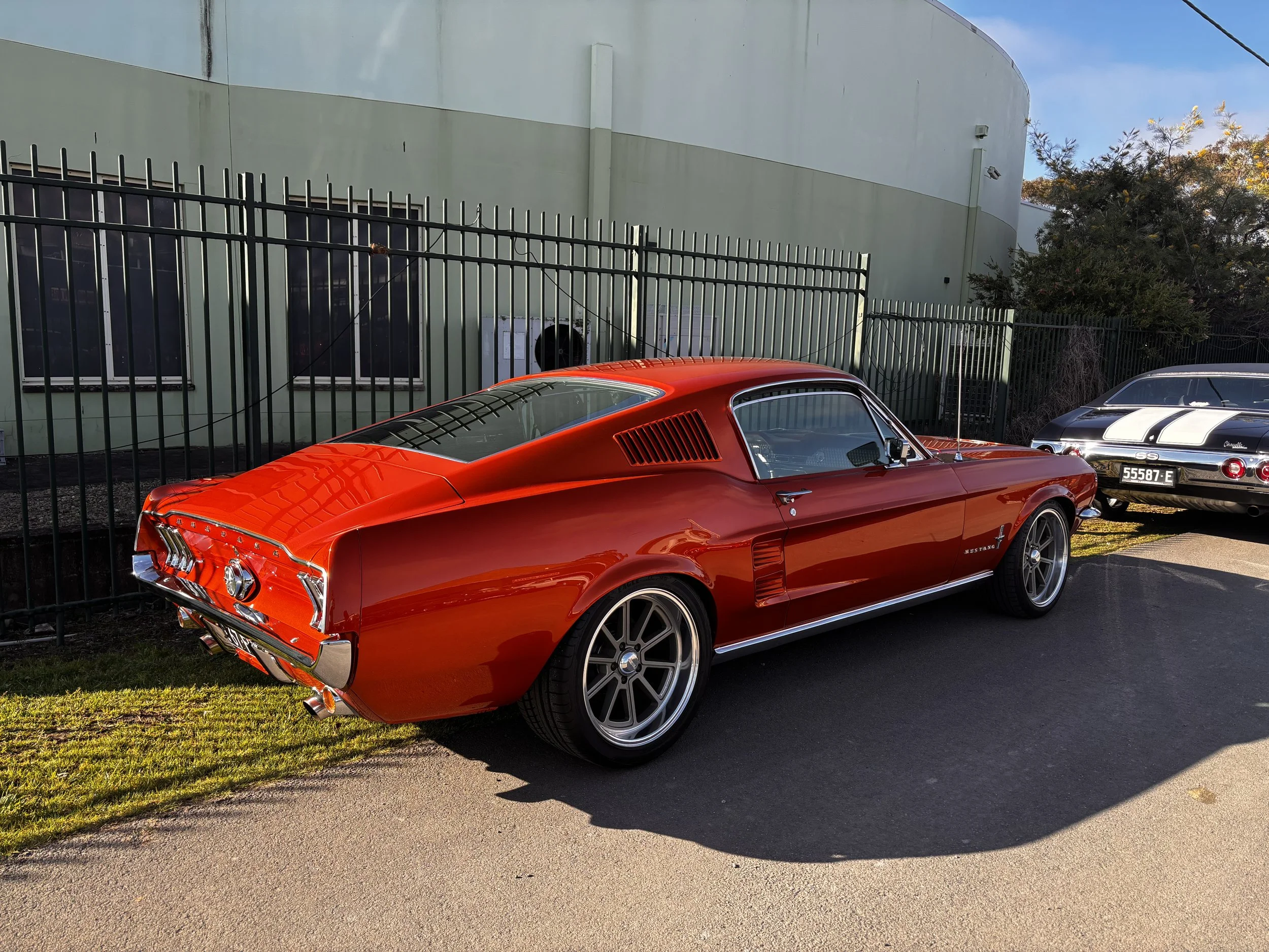 A red vintage Ford Mustang car parked beside a black metal fence and a pale green building, with another classic black car partially visible behind it, under a clear blue sky.