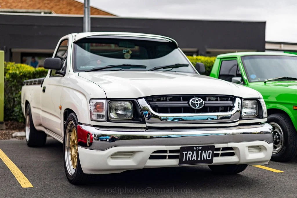 A white Toyota pickup truck with a custom license plate reading 'TRAINO,' parked in a parking lot next to a green vehicle. The truck has gold-colored wheels and chrome accents.