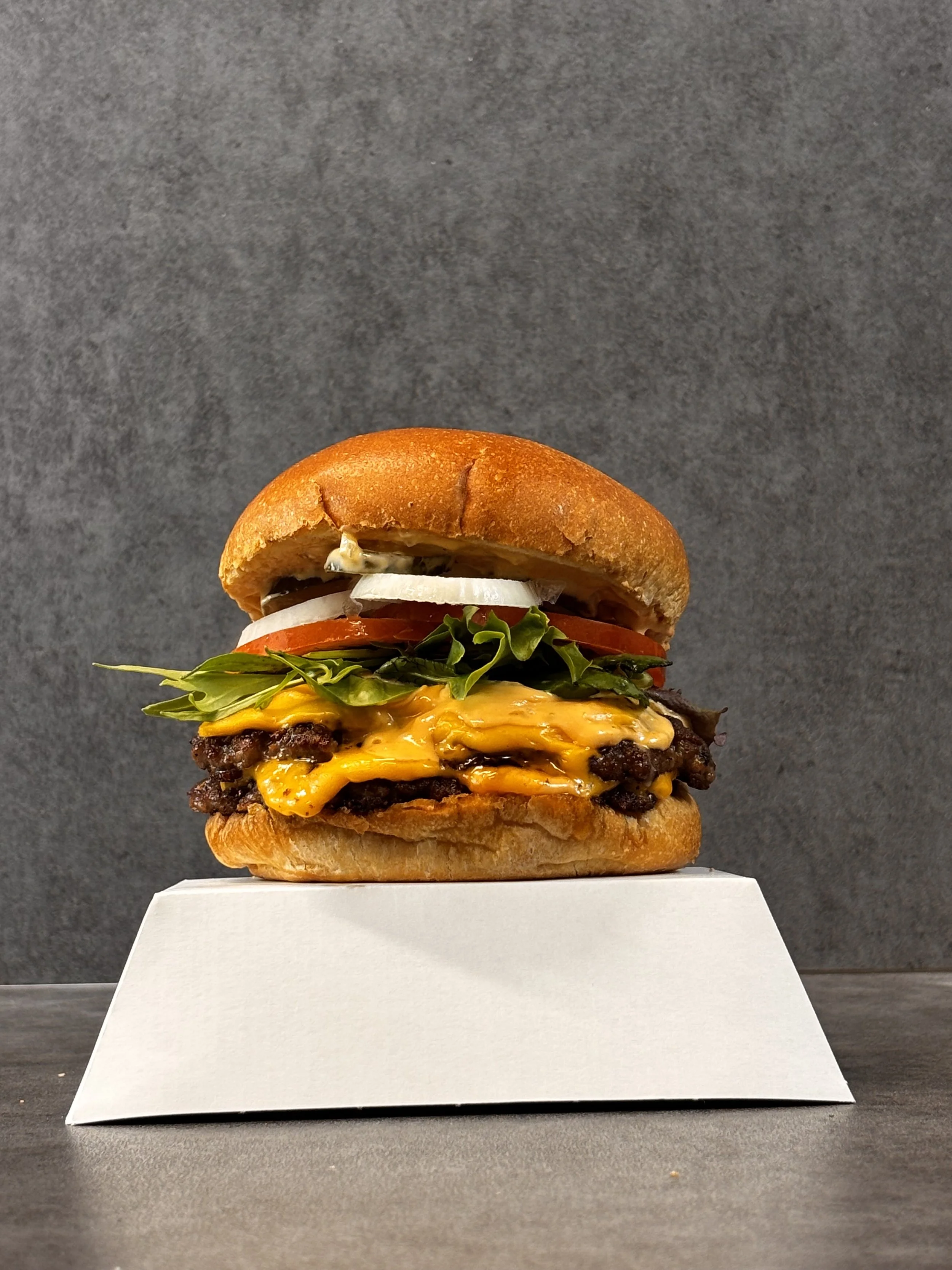 A cheeseburger with lettuce, tomato, onions, melted cheese, and a hamburger patty on a bun, placed on a white stand against a dark gray background.