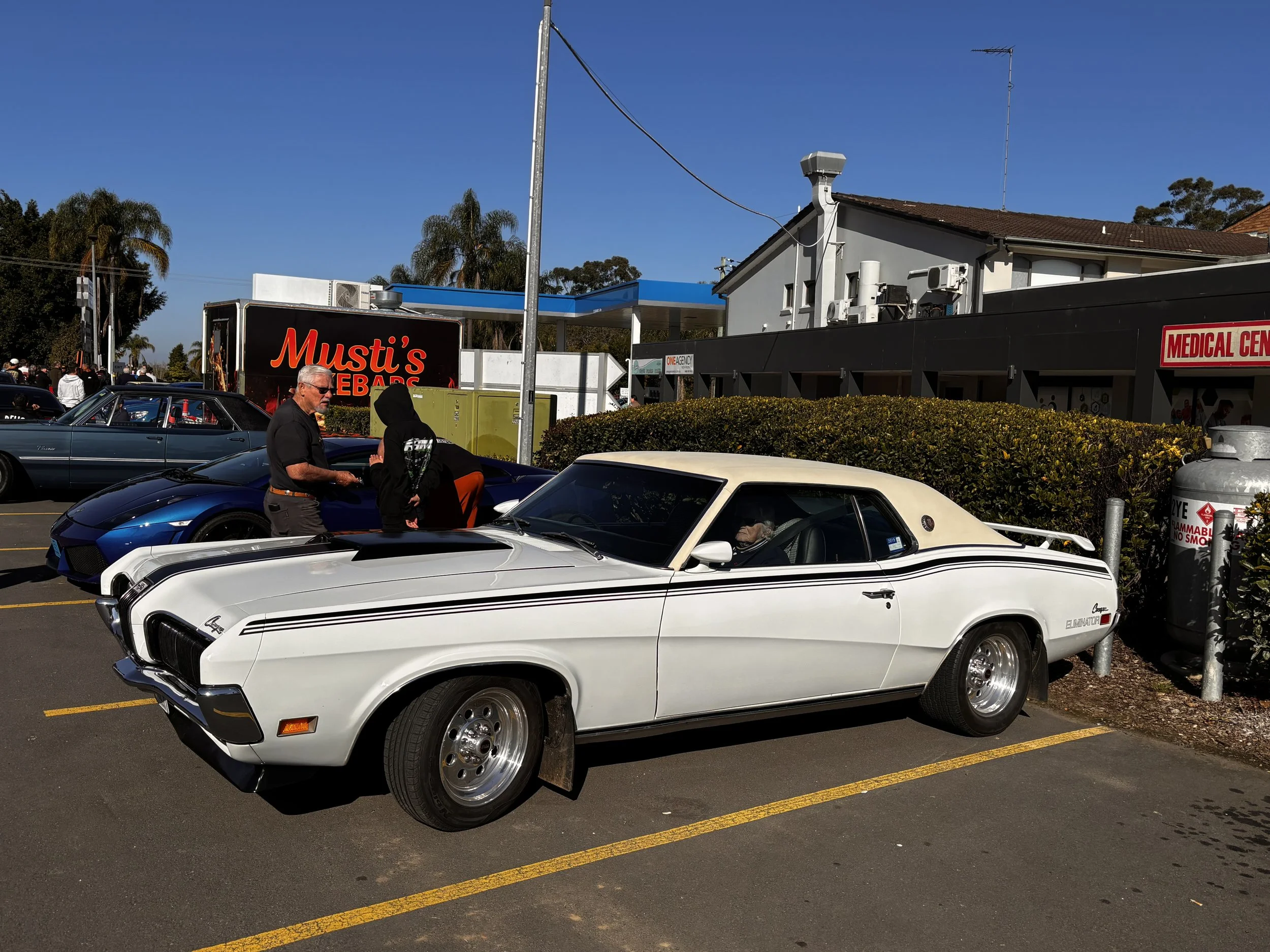 A parking lot filled with classic cars, including a white Pontiac Firebird, a blue Lamborghini Gallardo, and a black sports car. People are walking around, and there is a black food truck with red text that reads 'Musti's BBQ' in the background. The 