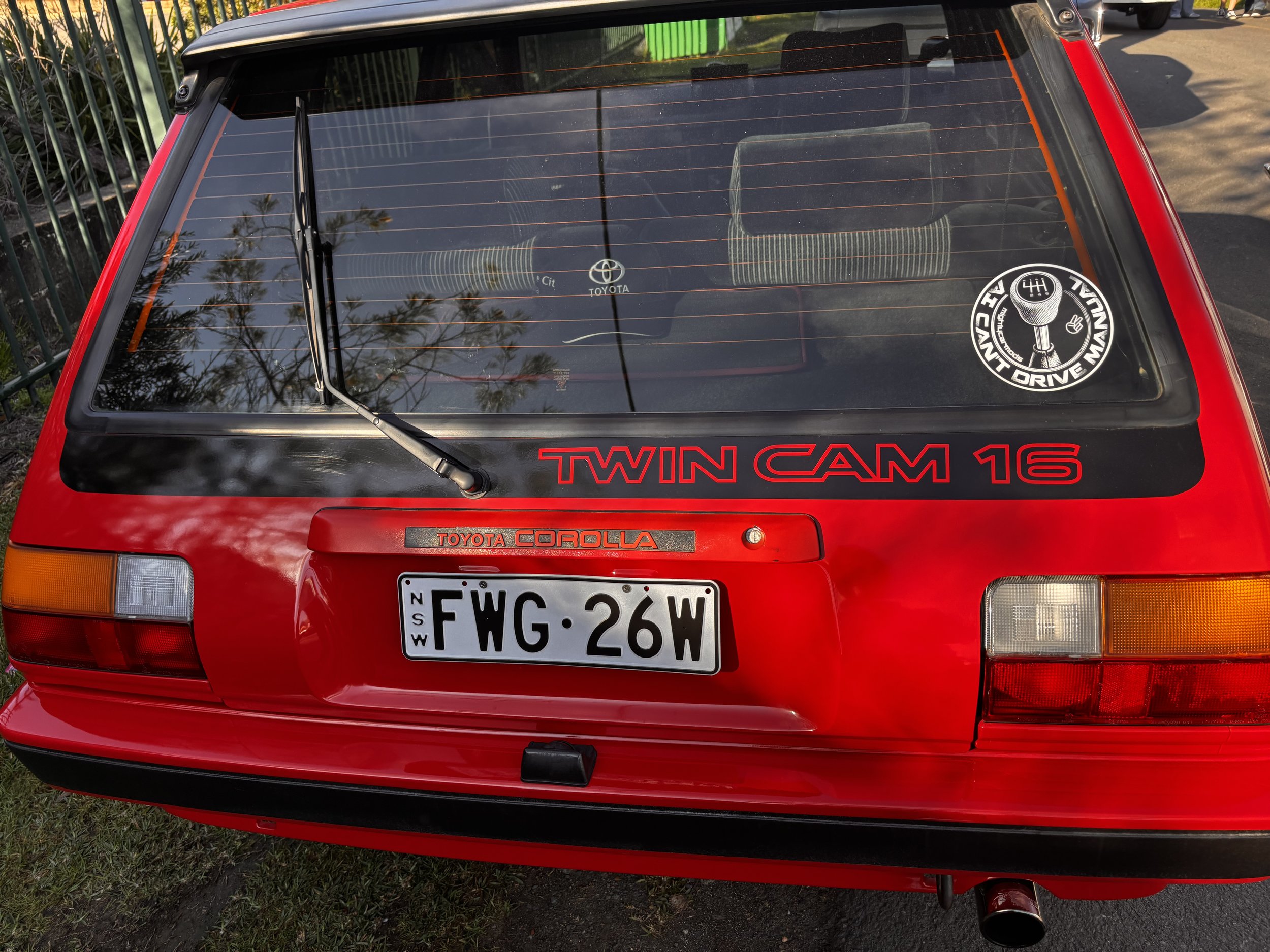The back of a red Toyota Corolla TWIN CAM 16 car with a European license plate reading 'FWG 26W'. The rear window has a sticker saying 'LONGLAST DRIVE'. The car is parked on grass near a gated fence, and reflections of trees are visible on the rear w