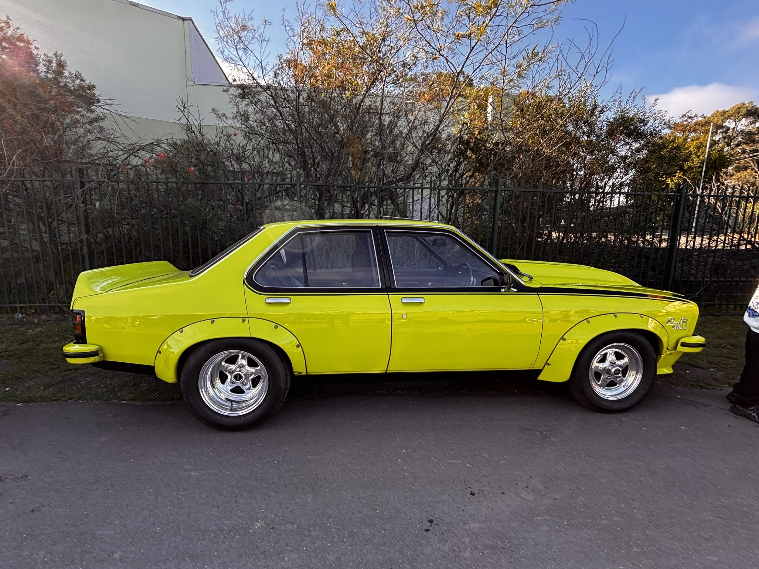 A bright yellow vintage car parked along a street with a black metal fence. Behind the fence are leafless trees with some yellow and red foliage. A modern light-colored building and trees are visible in the background against a partly cloudy sky.