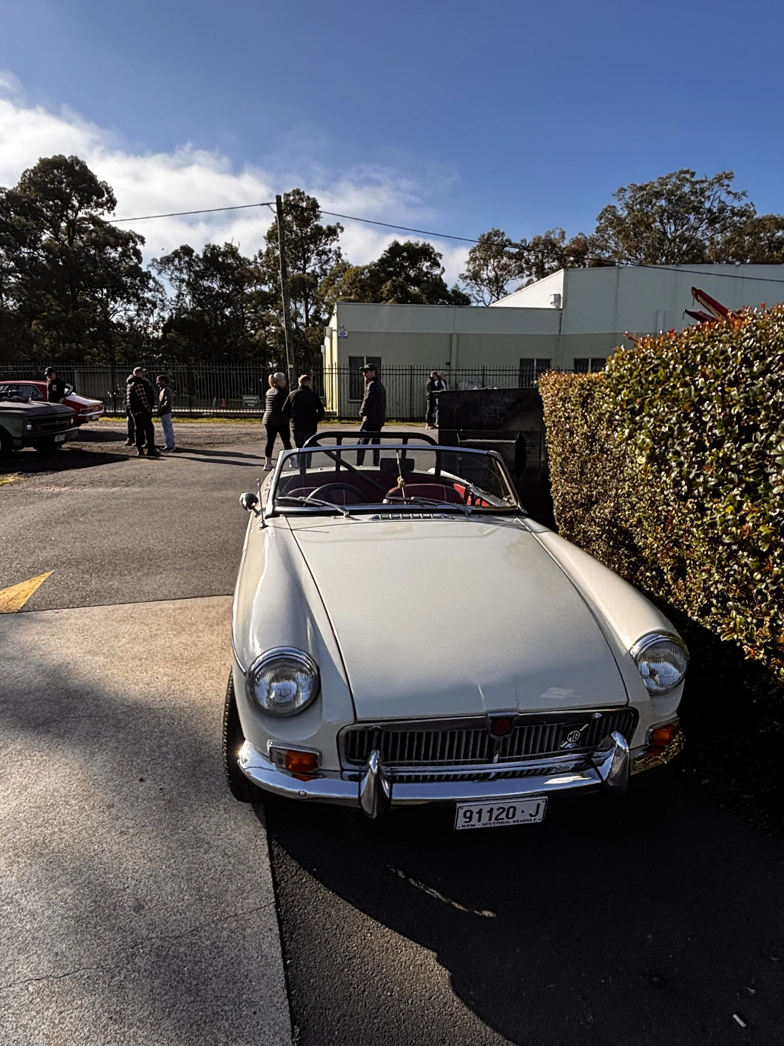 A vintage white convertible car parked in a parking lot with a group of people standing nearby, a building, trees, and a blue sky with some clouds in the background.