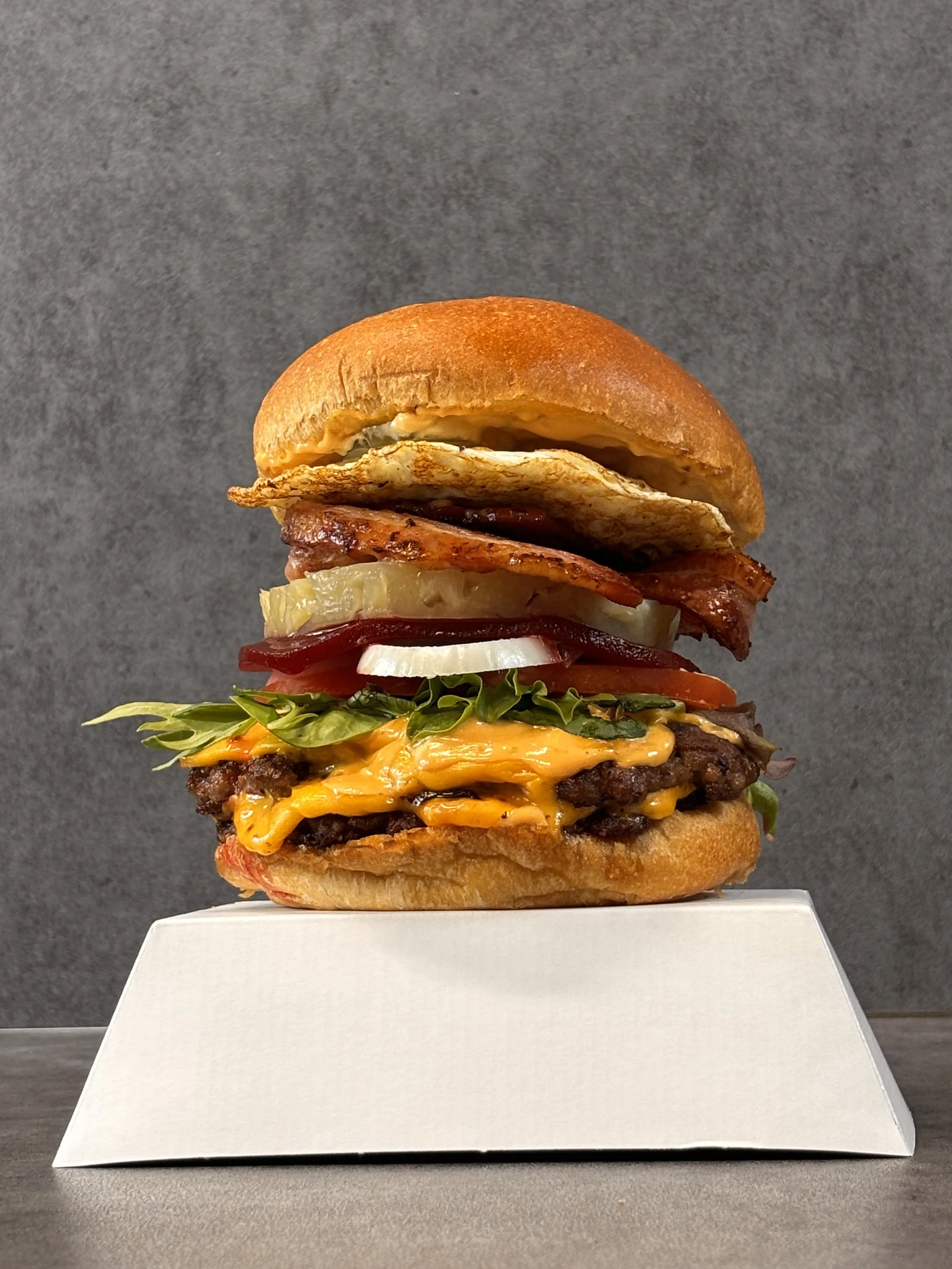 A tall burger with multiple layers including lettuce, tomato, onion, cheese, bacon, and a fried egg, all in a bun, served on a white box.