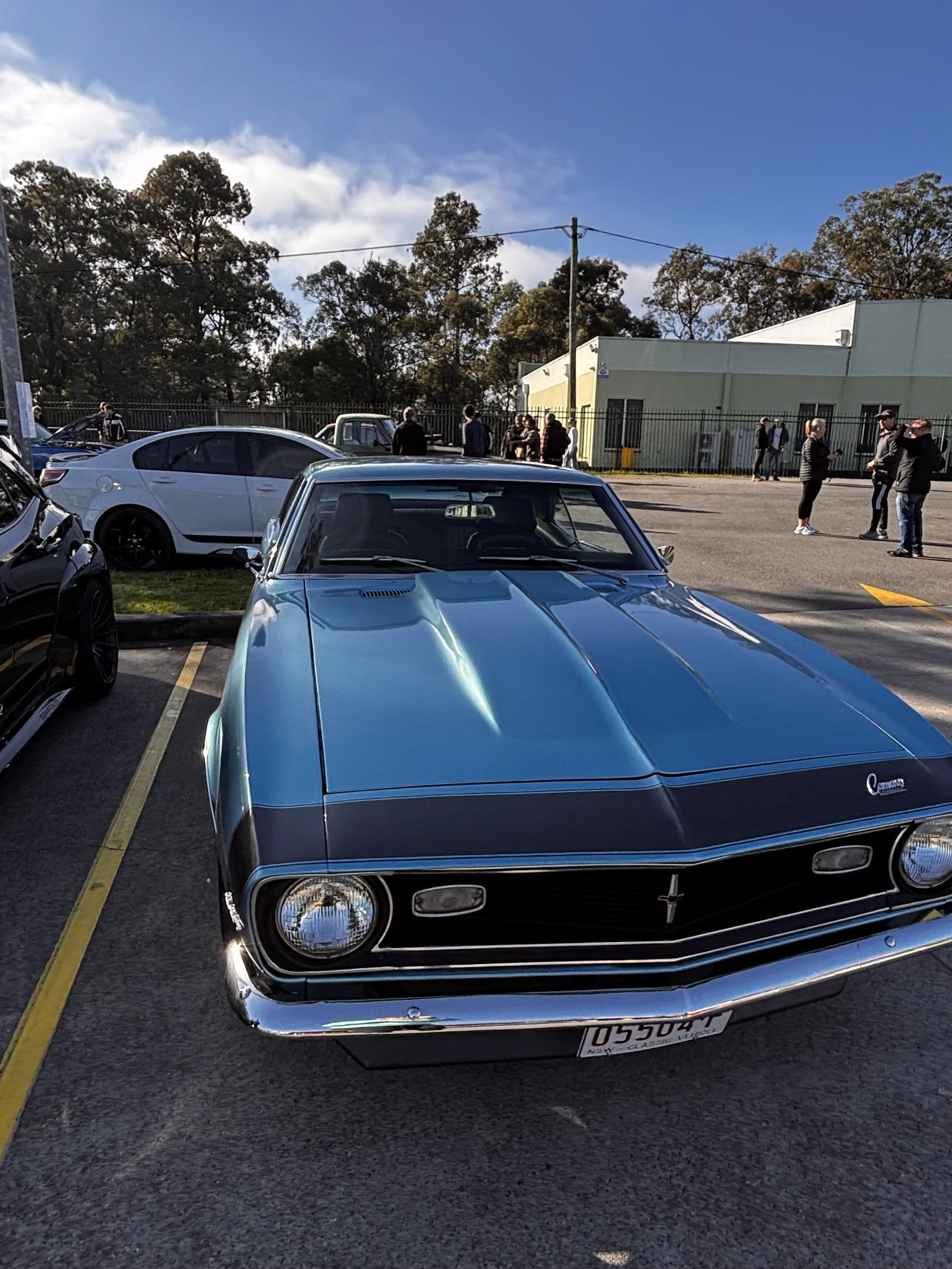 A parking lot with a classic blue Chevrolet Camaro in the foreground, other modern cars, and a group of people standing and talking near a fenced building with trees in the background on a sunny day.