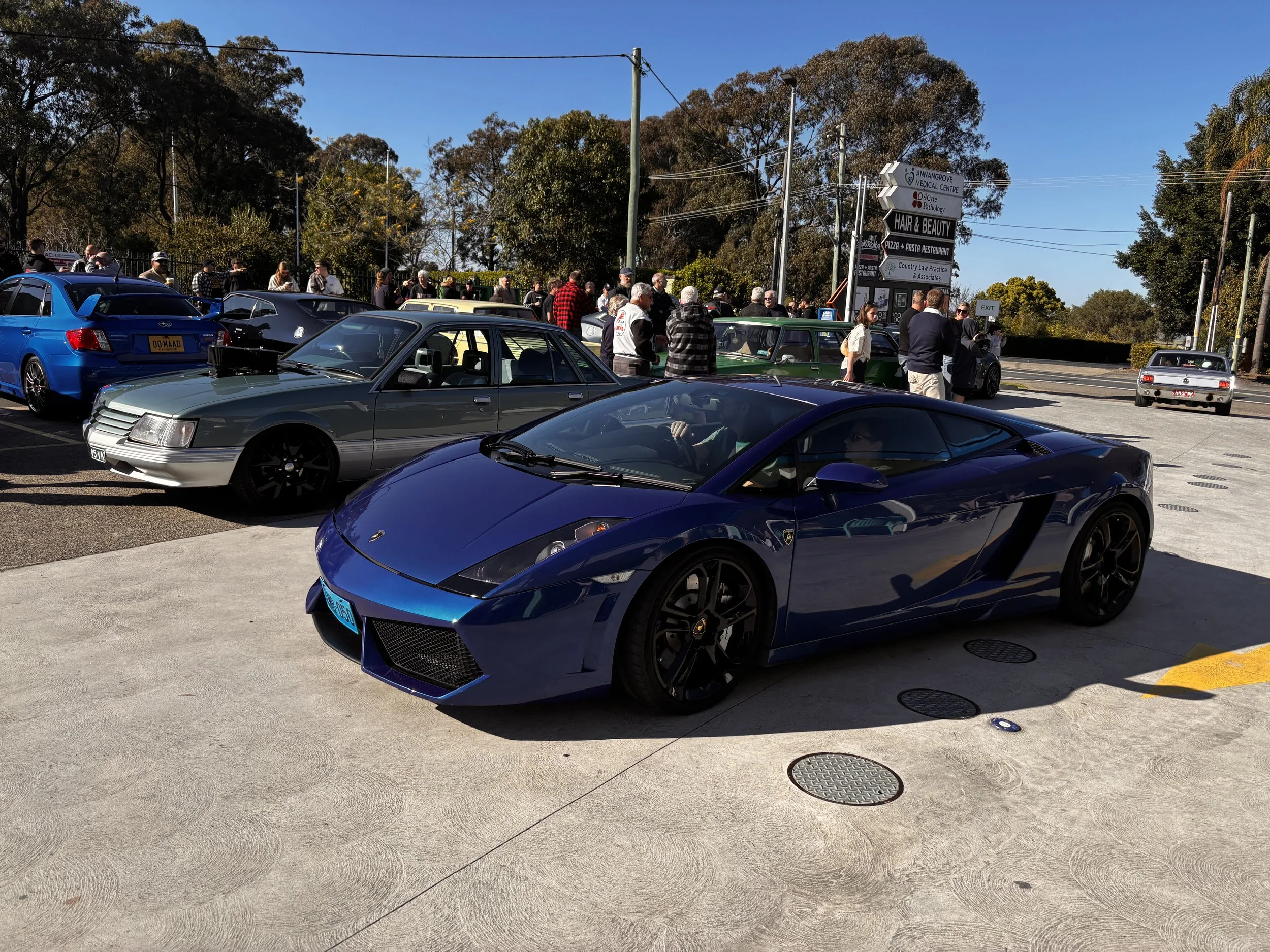 A parking lot filled with various cars and a group of people gathered near the cars, with trees and clear blue sky in the background and multiple directional signs.