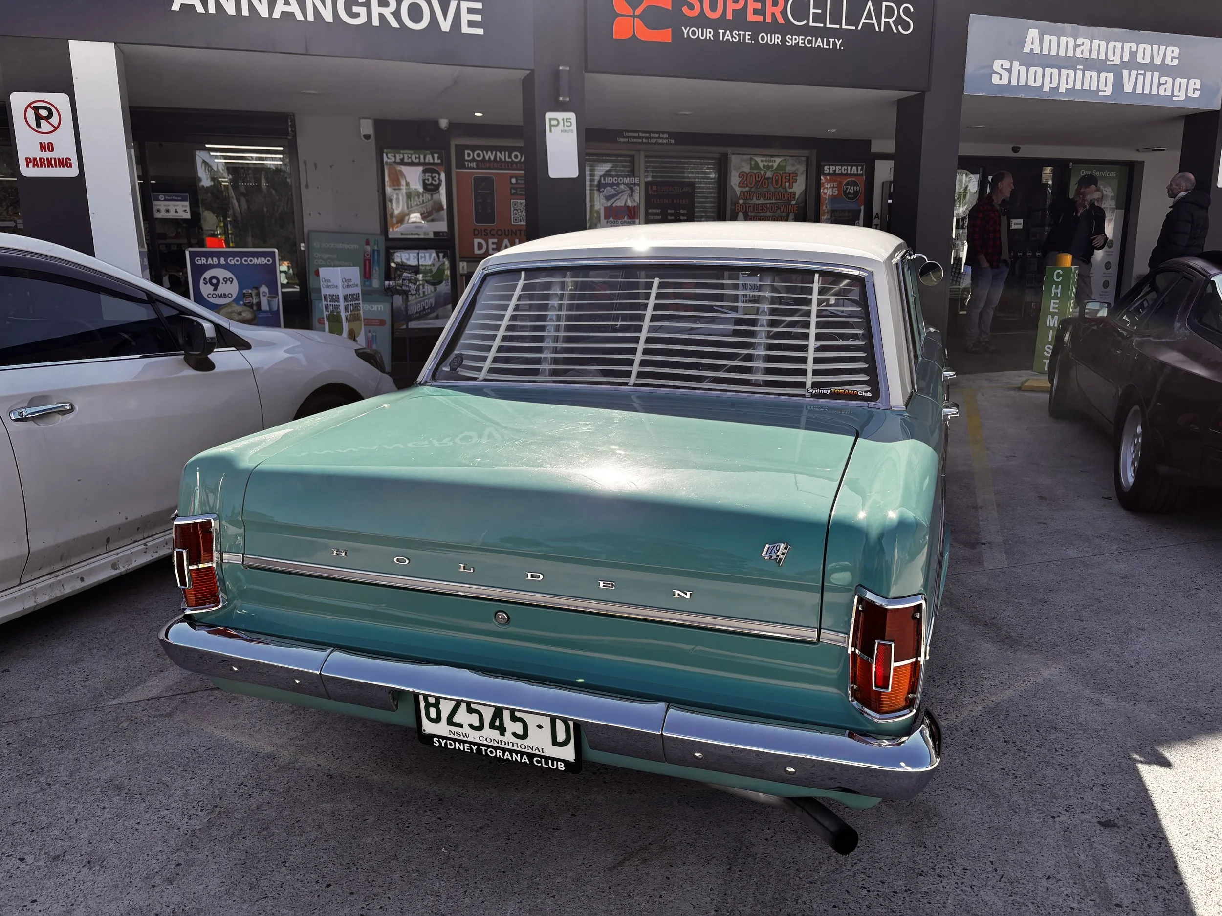 A vintage green Holden car with a white roof and license plate 82545 D parked in front of a shopping center entrance, with people standing nearby. The shopping center has signs for Super Cellars and Annangrove Shopping Village.