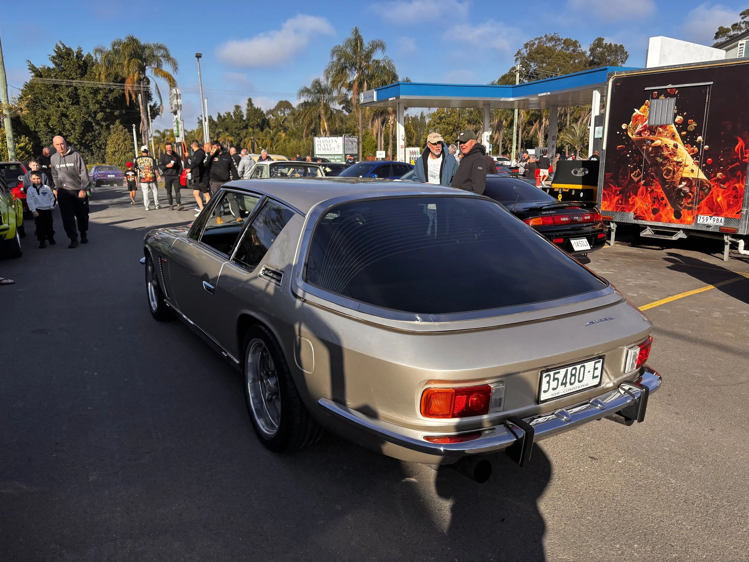 A parking lot filled with classic cars and people. There is a food truck with a pizza design on the side, trees, and a blue sky in the background.