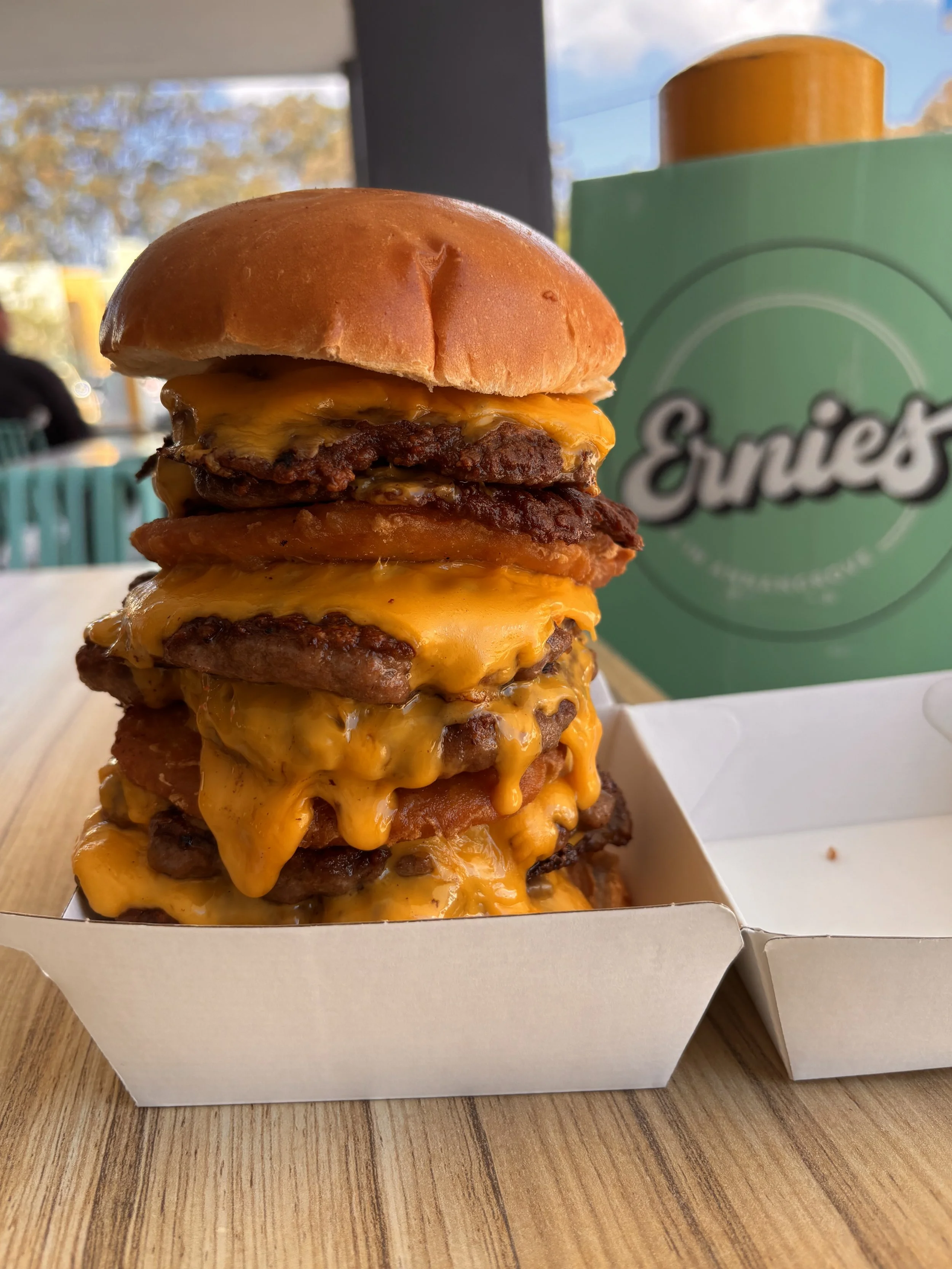 Close-up of a stacked cheeseburger with multiple beef patties and melted cheese in a white takeout box on a wooden table.