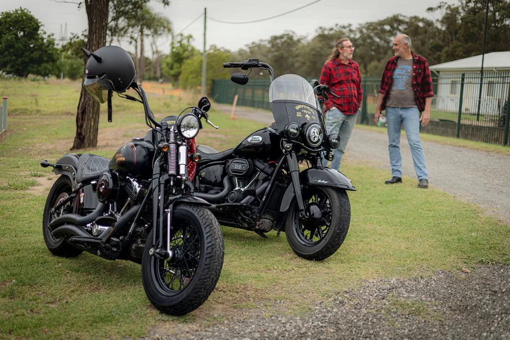 A black motorcycle with a helmet hanging on the handlebar parked on grass, with two men in plaid shirts having a conversation in the background near a fence and a building.