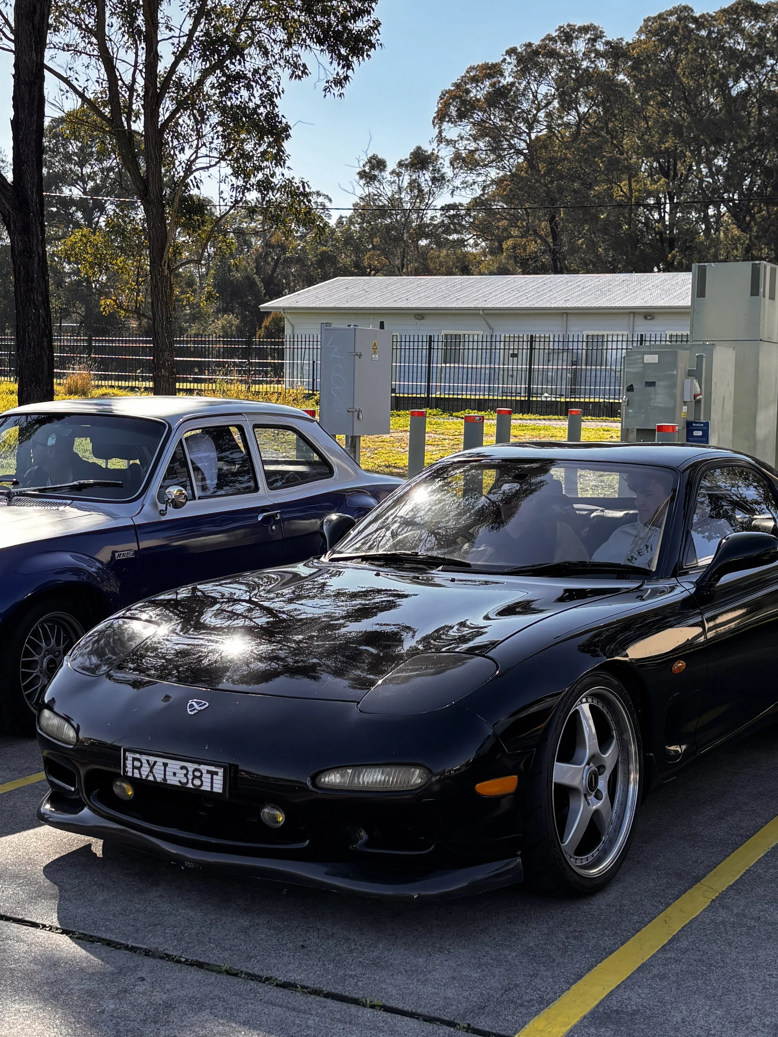 A parking lot with two black sports cars, one with a visible license plate, parked side by side in front of a fenced area with trees and a small white building with a metal roof. The sky is clear and sunny.
