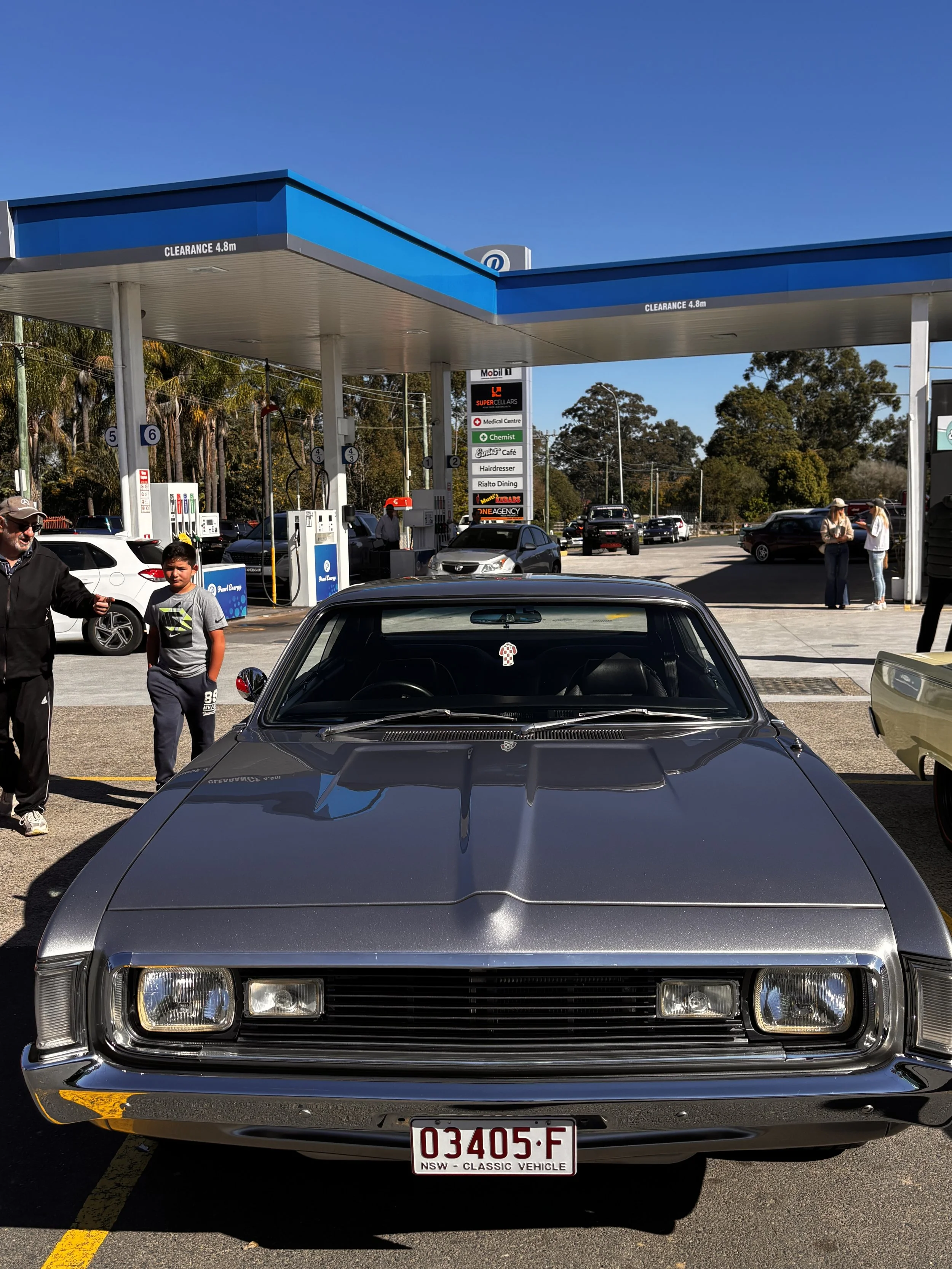 Front view of a gray classic car with Australian license plate, parked at a gas station with people nearby and trees in the background.