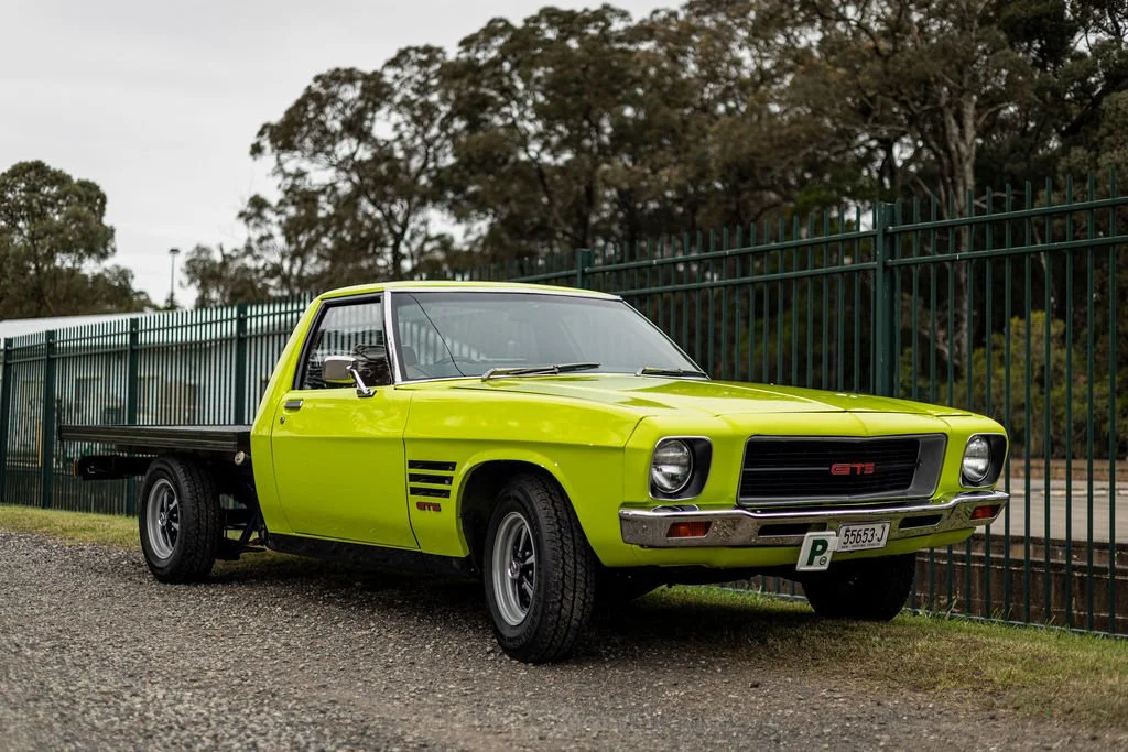 A bright green vintage pickup truck parked next to a green metal fence on a gravel surface, with trees in the background.