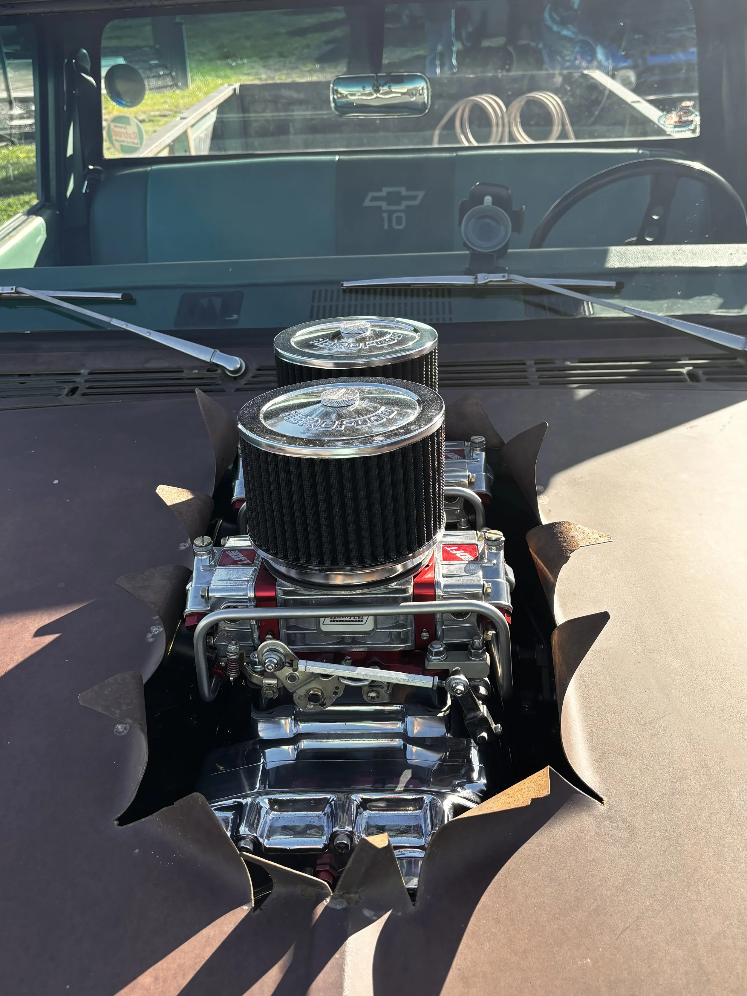 View inside a vintage Chevrolet truck with a high-performance engine with dual round air filters, seen through the windshield. The truck's dashboard has a Chevrolet emblem, with chrome and black details, and two white wiper blades are visible resting
