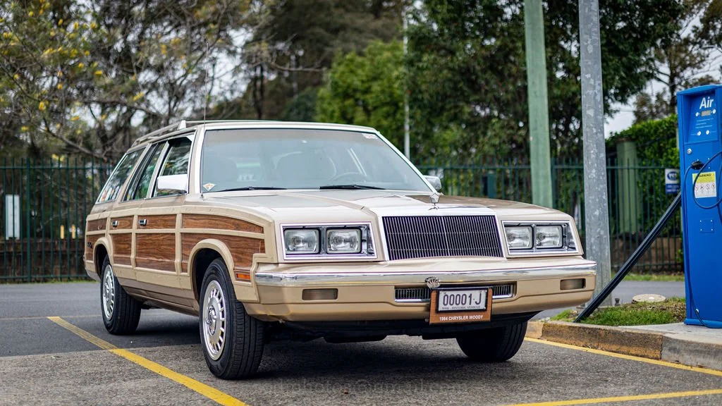 A vintage 1984 Chrysler Woodoy station wagon parked at a charging station.
