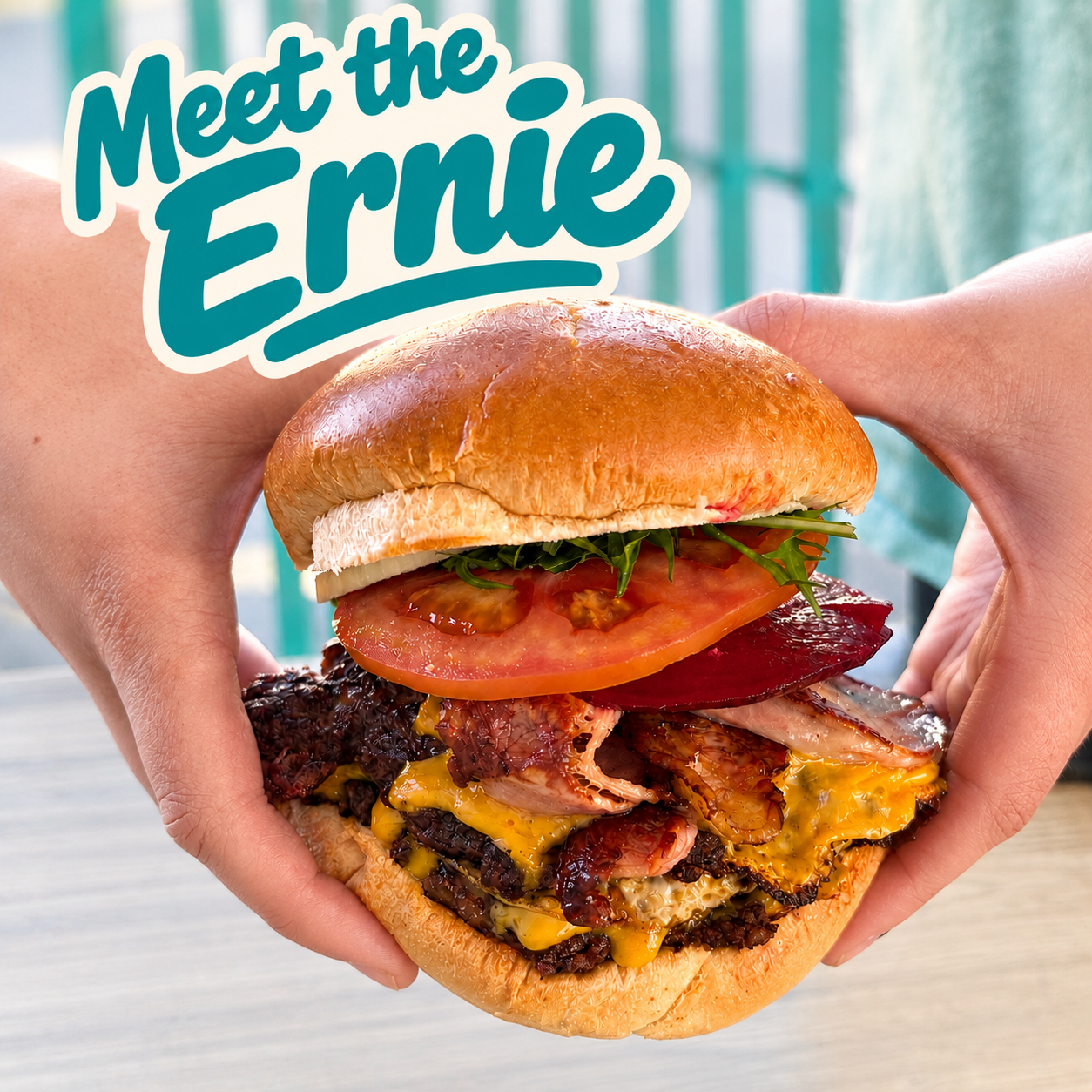 A person holding a large burger with various ingredients and a bun labeled "Meet the Ernie" in a bright setting.