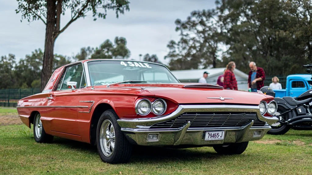A vintage red car parked on the grass at a car show, with a 'For Sale' sign on the windshield, surrounded by trees and other vehicles.