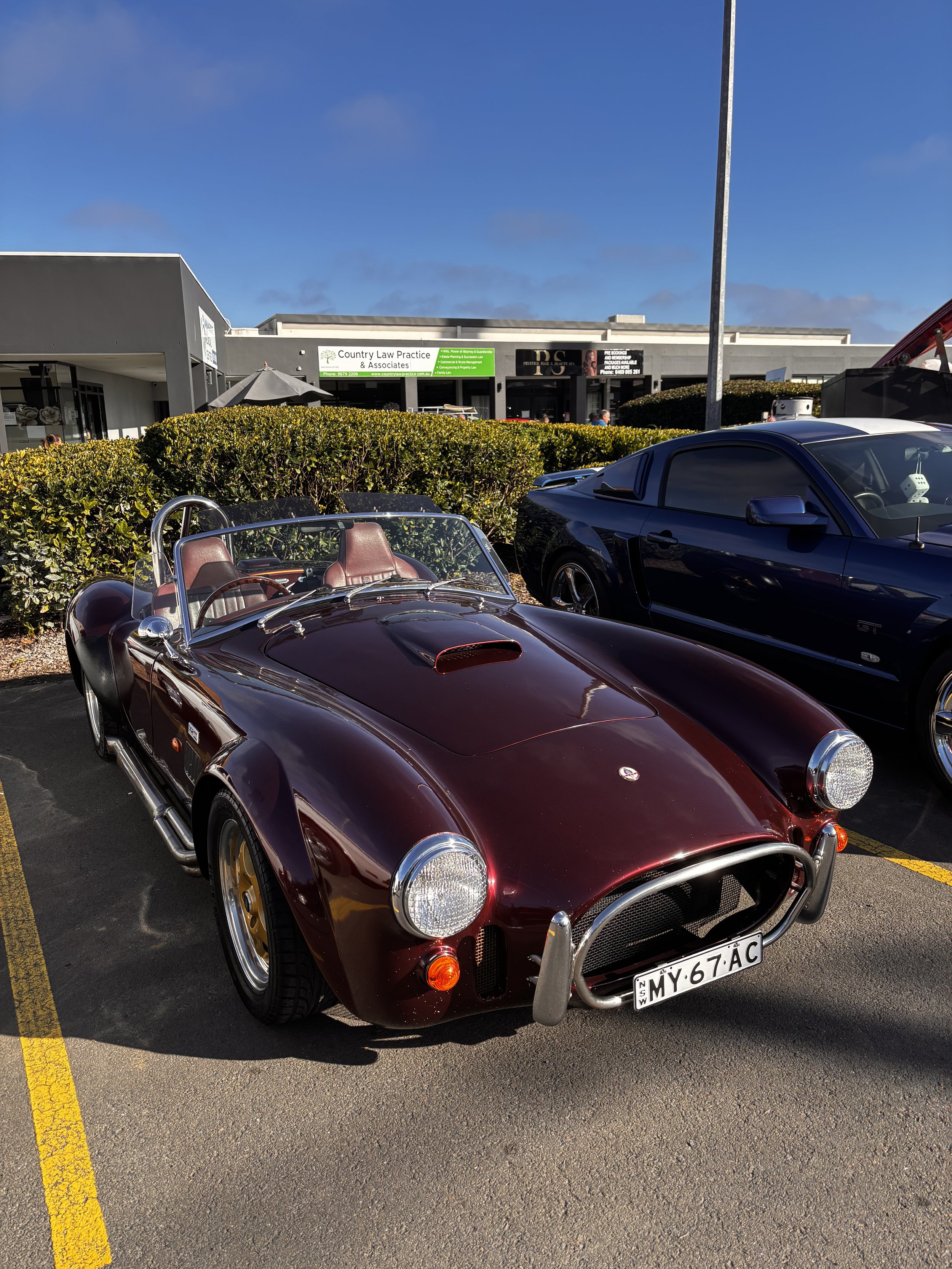 A classic maroon convertible sports car parked in a parking lot with two modern black cars nearby. The background features a shopping plaza with a hedge, a sign for a law practice, and a blue sky.