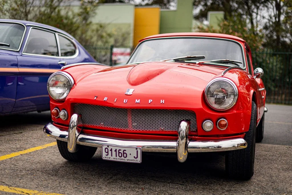 Red vintage Triumph sports car parked next to a blue vintage car in a parking lot.