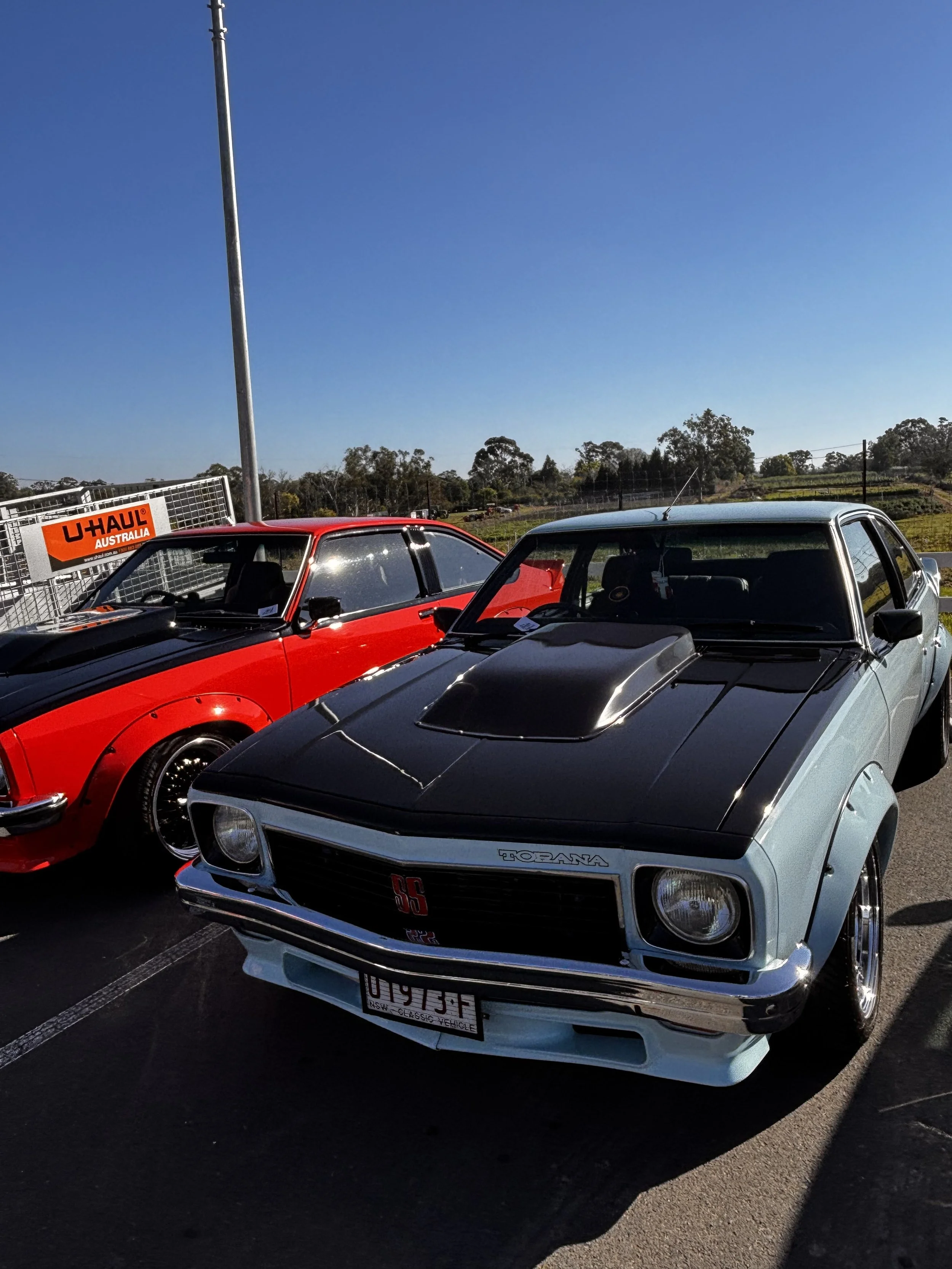 Close-up of two classic sports cars parked side by side in an outdoor parking lot with a clear blue sky and distant trees in the background.