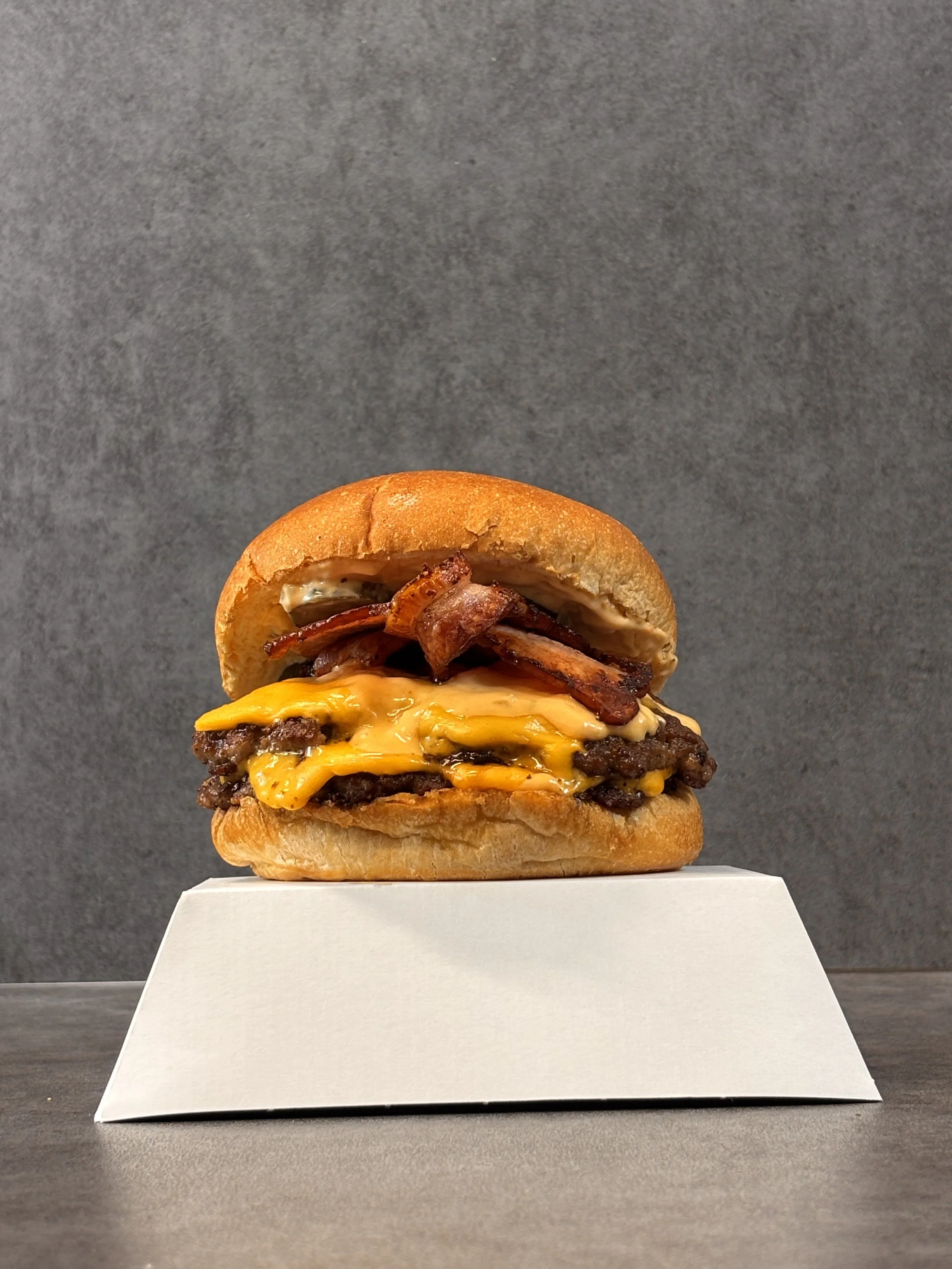 A cheeseburger with bacon on a bun, served on a white platform with a gray background.