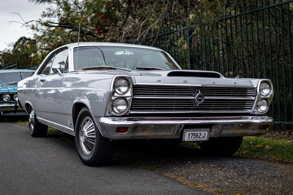 A vintage silver Ford car with a prominent grille and hood scoop, parked on a street next to a fence.