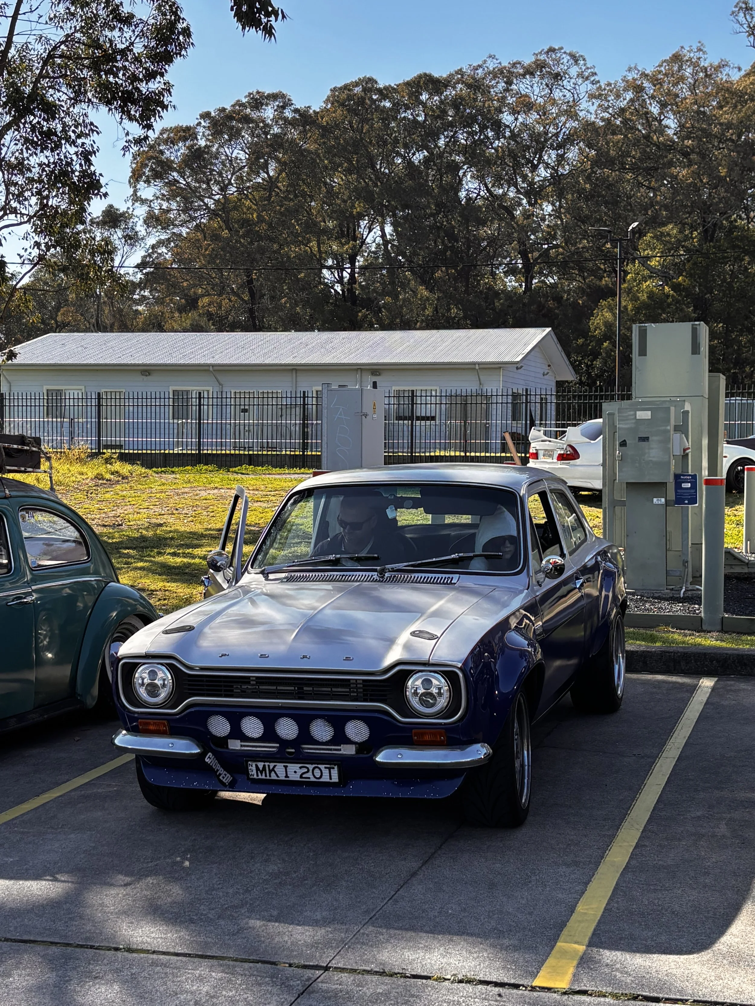 A parking lot with a classic dark blue Ford Maverick car in the foreground, facing forward, with a white building and trees in the background. There are other parked cars and electrical equipment nearby.