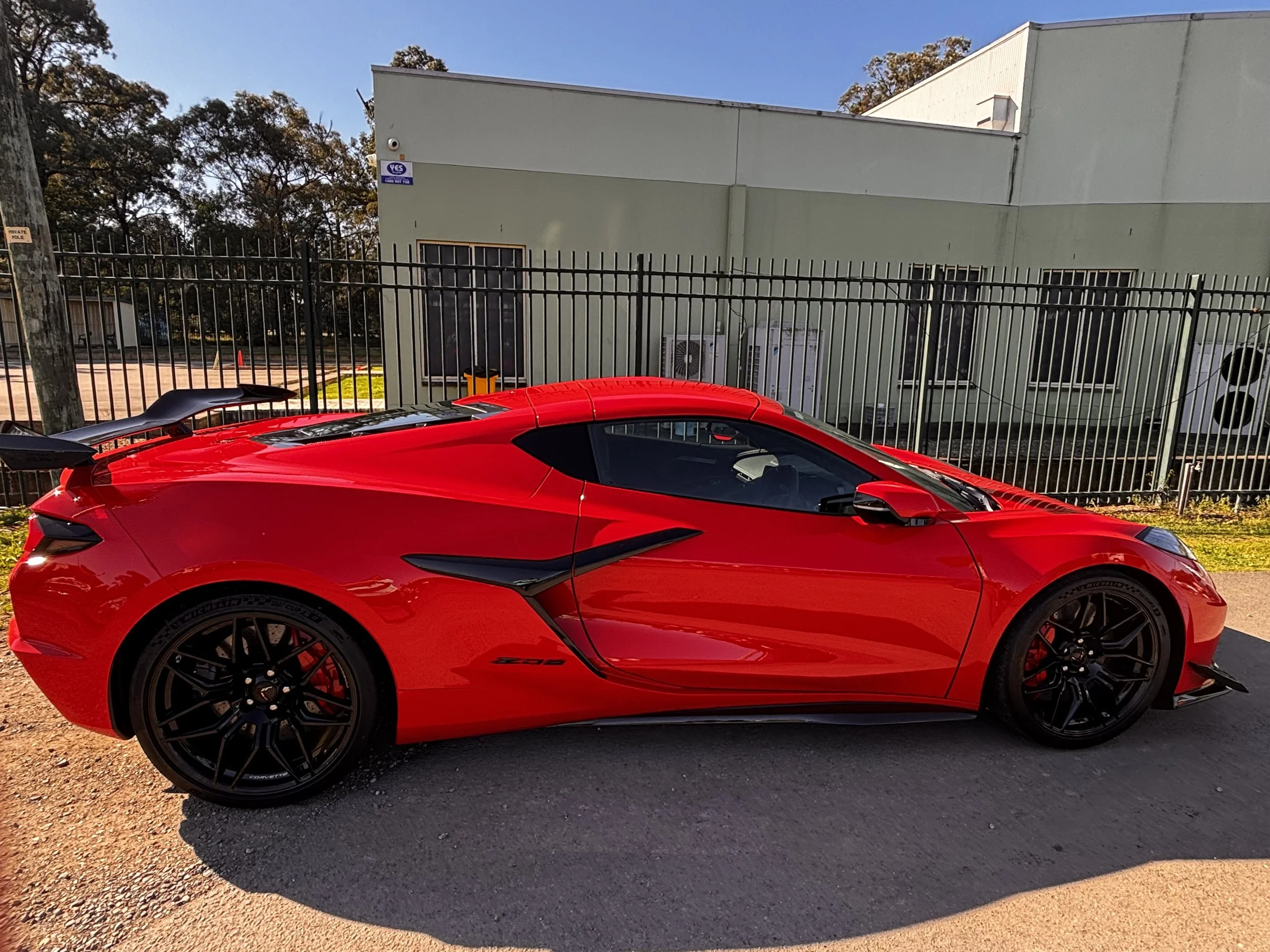 A bright red sports car parked on a gravel surface in front of a black metal fence and a light green building with air conditioning units. Trees and a clear blue sky are visible in the background.