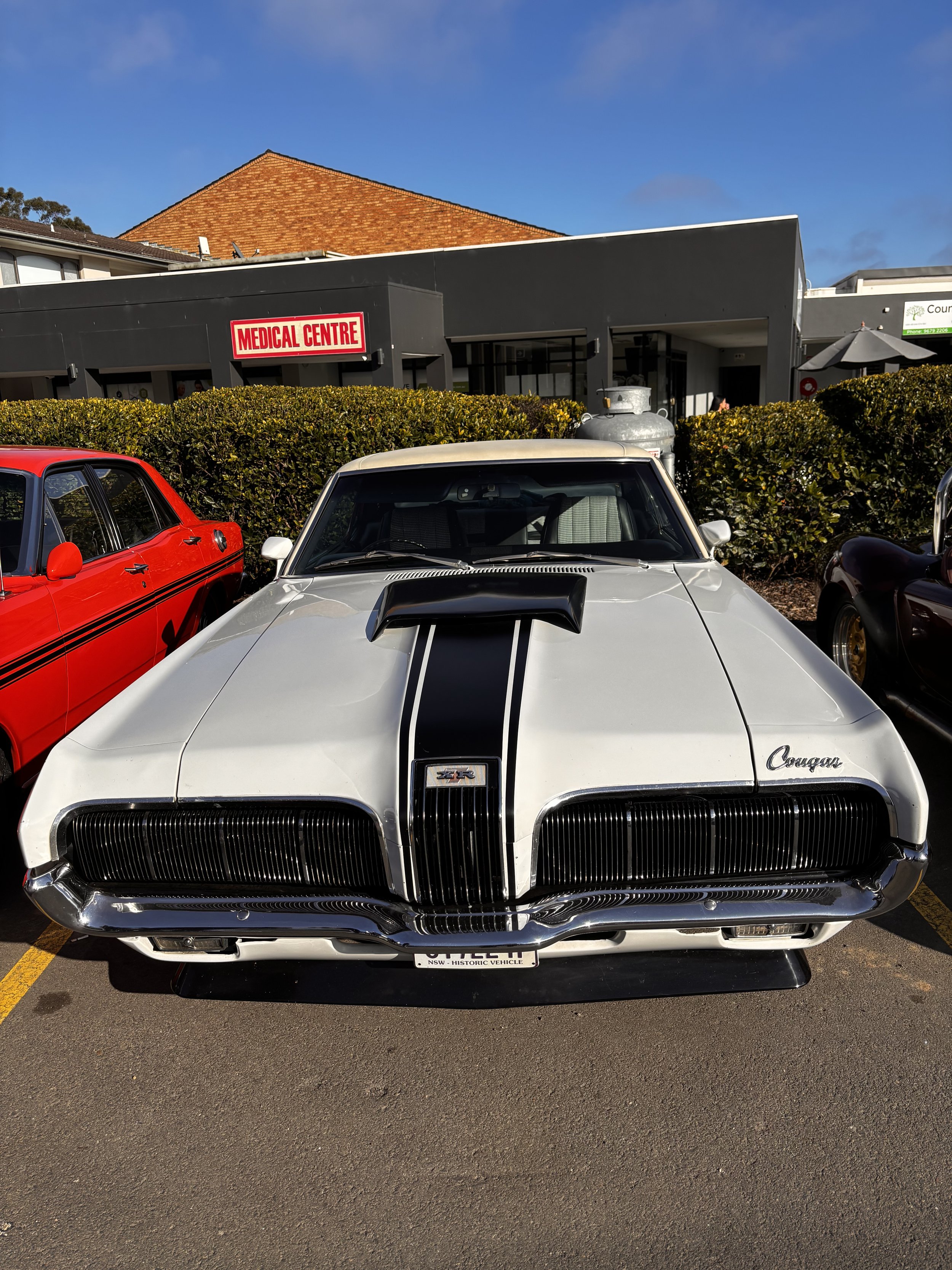 Front view of a white classic muscle car with black racing stripes, parked in a lot with other cars visible on sides, with a black building labeled 'Medical Centre' and a hedge in the background, under a blue sky.
