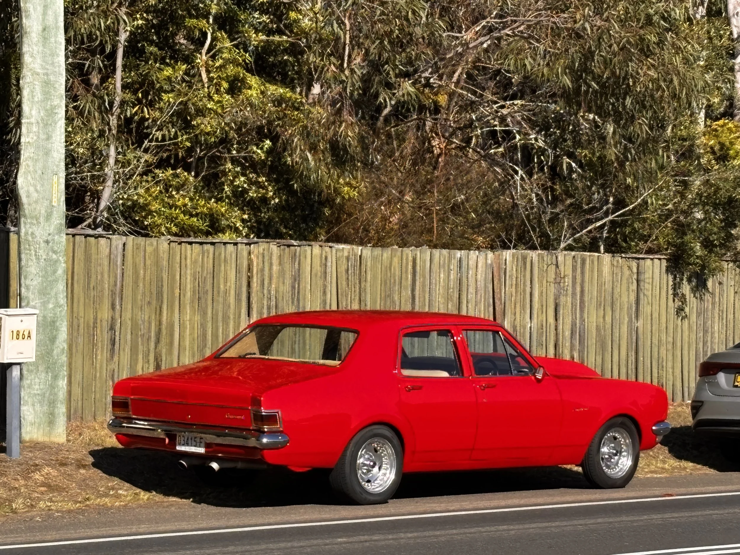 Red vintage car parked beside a wooden fence with trees and utility pole in the background.