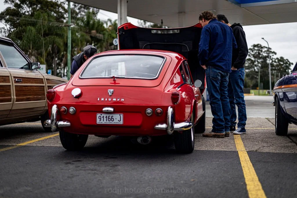 Group of three people examining the engine of a red vintage Triumph sports car parked in a parking lot on a cloudy day.